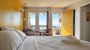 Hotellihuone, jossa on sänky, pieni pöytä, kaksi tuolia, parveke, jolta on näkymä, ja ovi, joka johtaa toiseen huoneeseen.