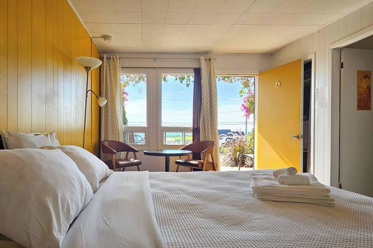 Habitación de hotel con cama, una mesita, dos sillas, un balcón con vistas y una puerta que da a otra habitación.