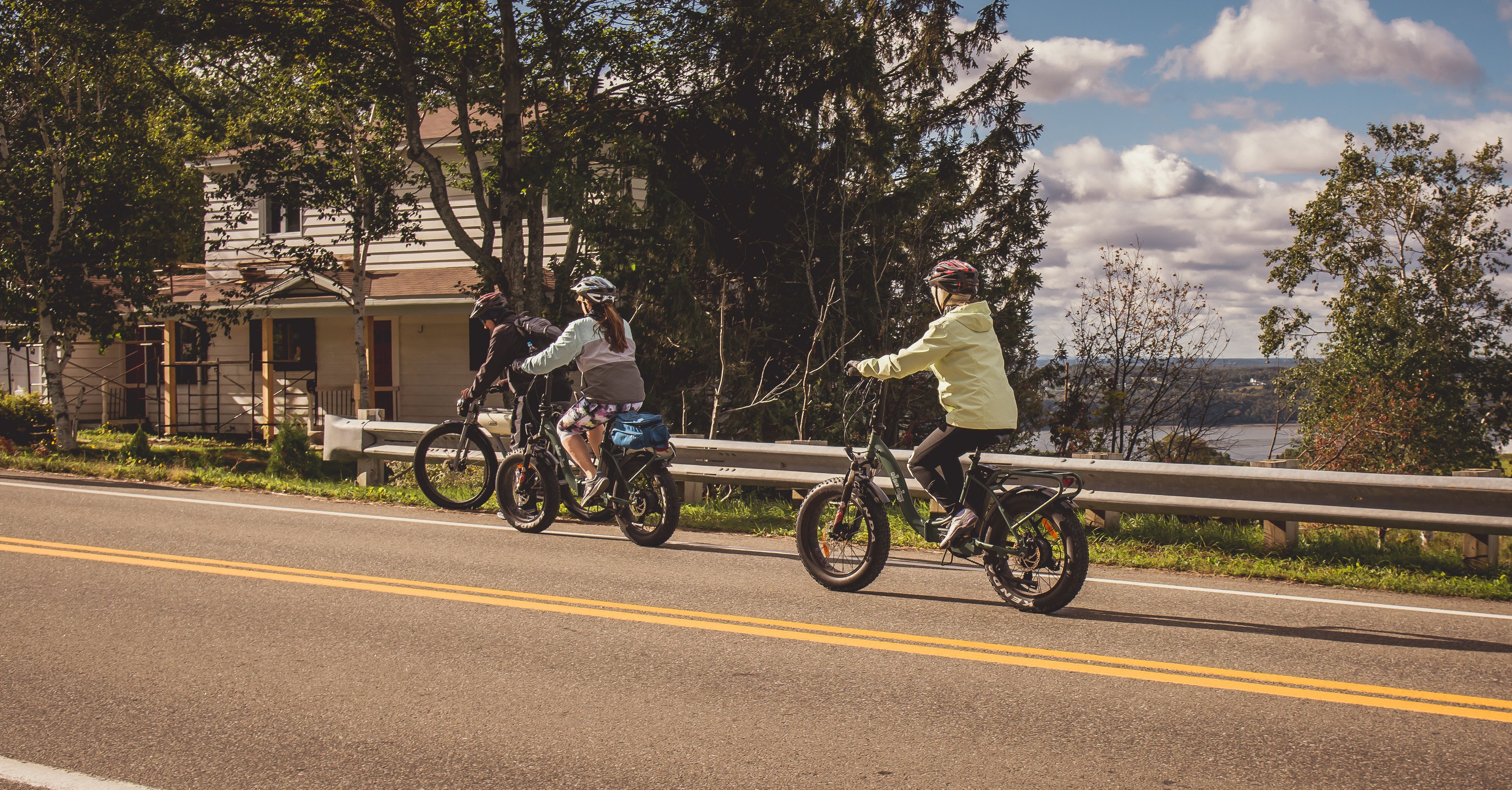 Three cyclists on a road with a guardrail, trees, and a building in the background.