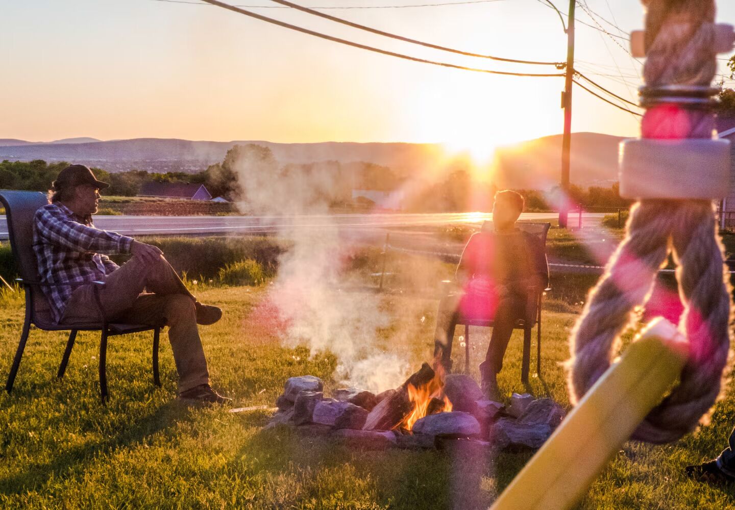 Two people sitting by a campfire in a grassy field with a sunset in the background.