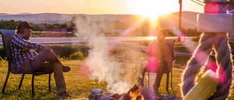Two people sitting by a campfire in a grassy field with a sunset in the background.