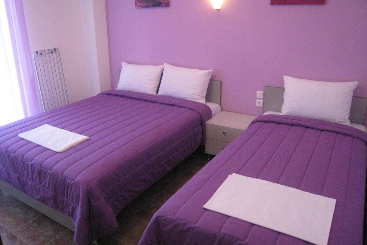 Habitación doble con dos camas, cada una con cubrecama morado y almohadas blancas. Cada cama tiene una mesita de noche con una lámpara.