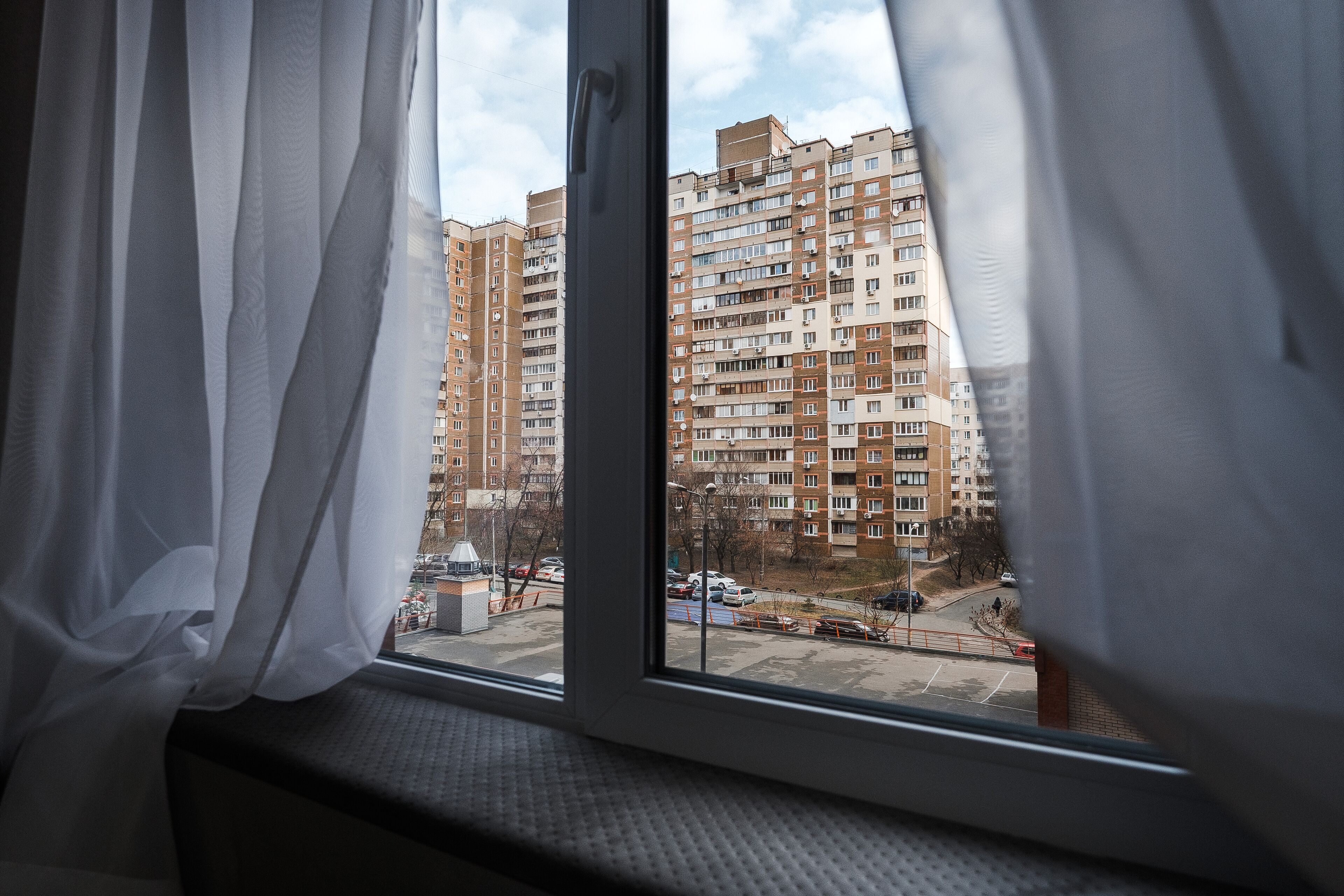 A window with white curtains, a view of a multi-story residential building, and a parking area with cars and construction equipment.