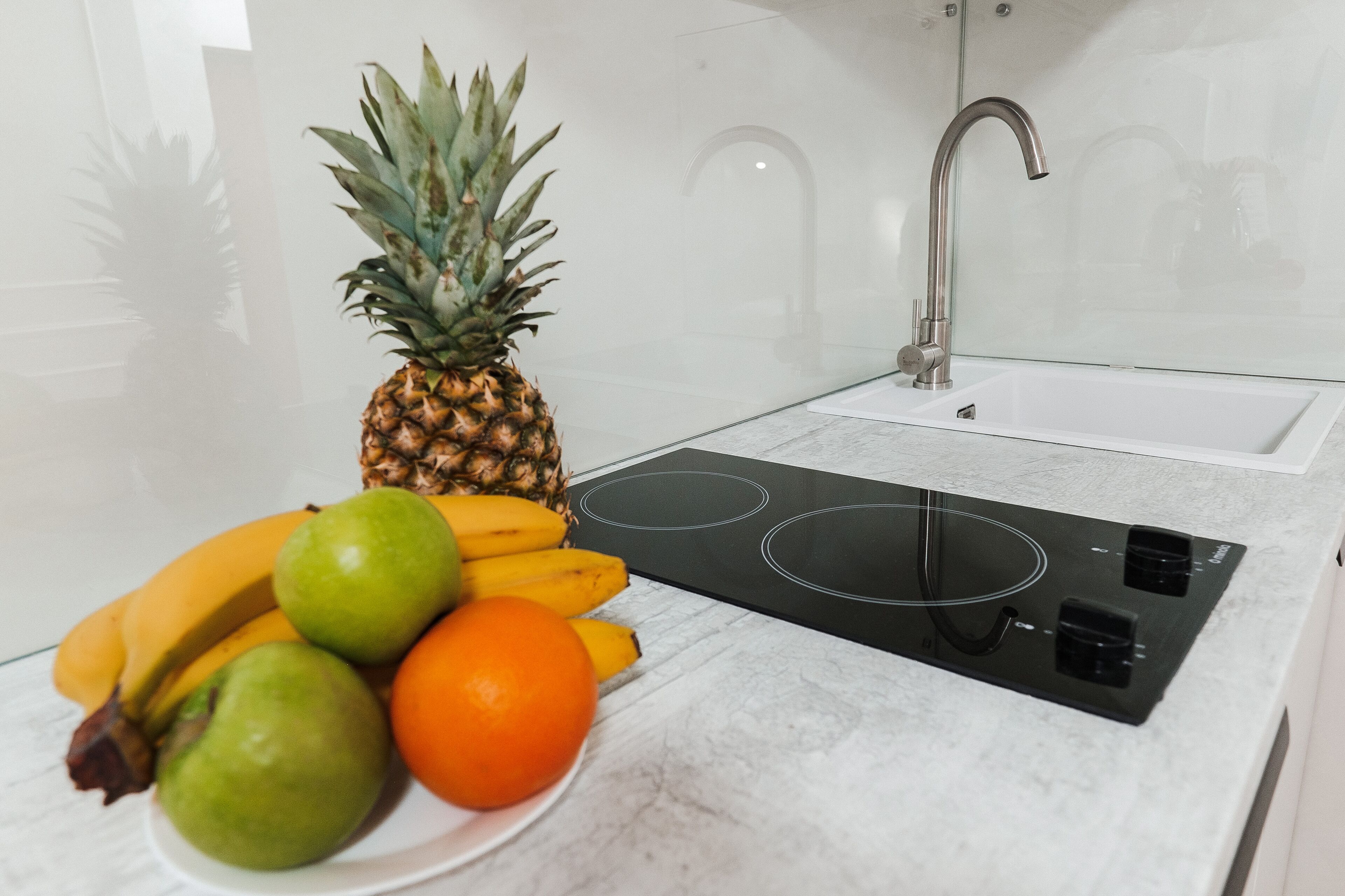 Una cucina con un piano di lavoro bianco, un piano cottura a induzione nero, un rubinetto in acciaio inossidabile e una ciotola di frutta contenente un'ananas, banane e arance.