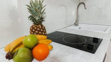 Una cucina con un piano di lavoro bianco, un piano cottura a induzione nero, un rubinetto in acciaio inossidabile e una ciotola di frutta contenente un'ananas, banane e arance.