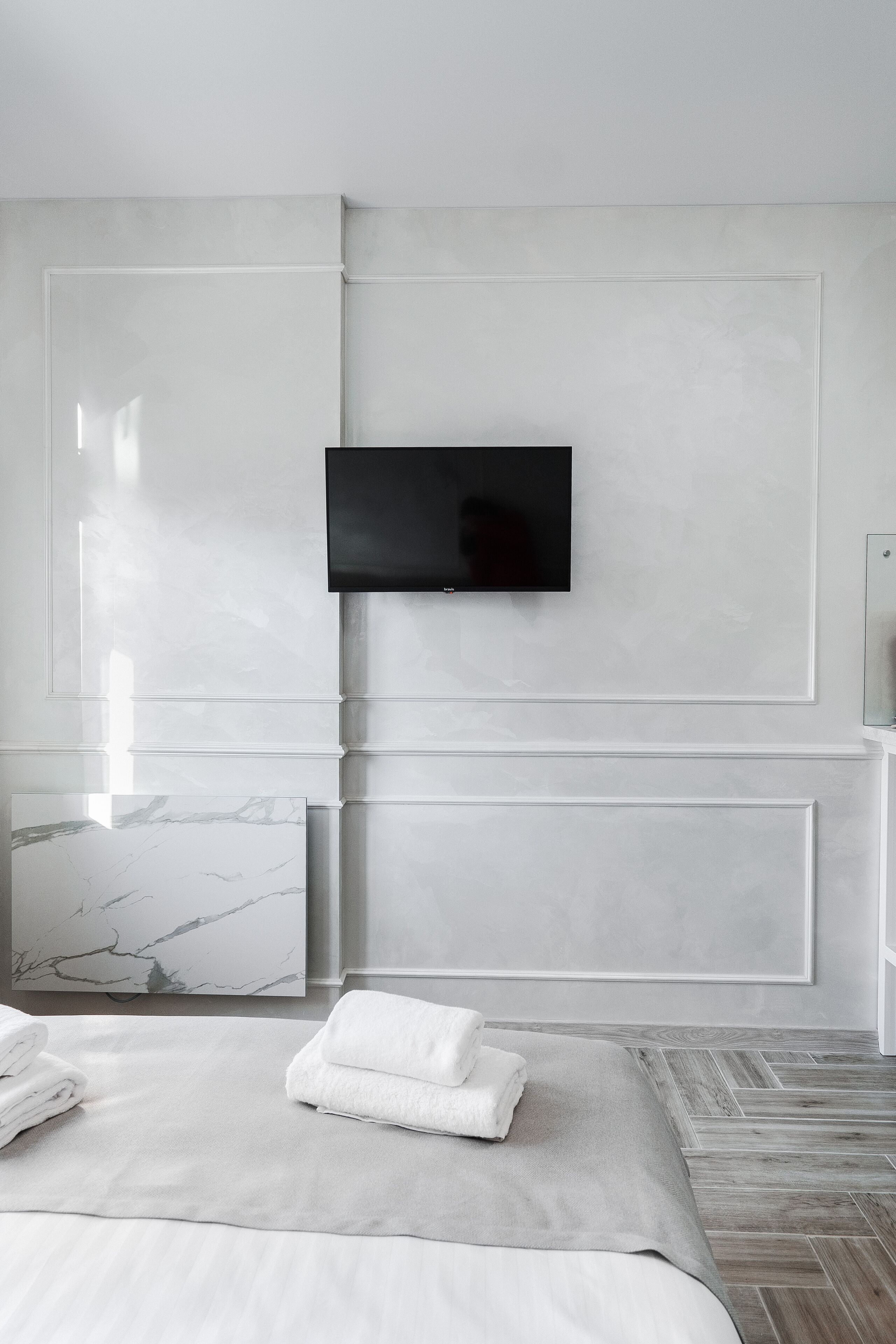 Quarto de hotel moderno com televisão de tela plana na parede, painel de parede em mármore, cama bem arrumada com roupa de cama branca e piso de madeira com padrão herringbone.