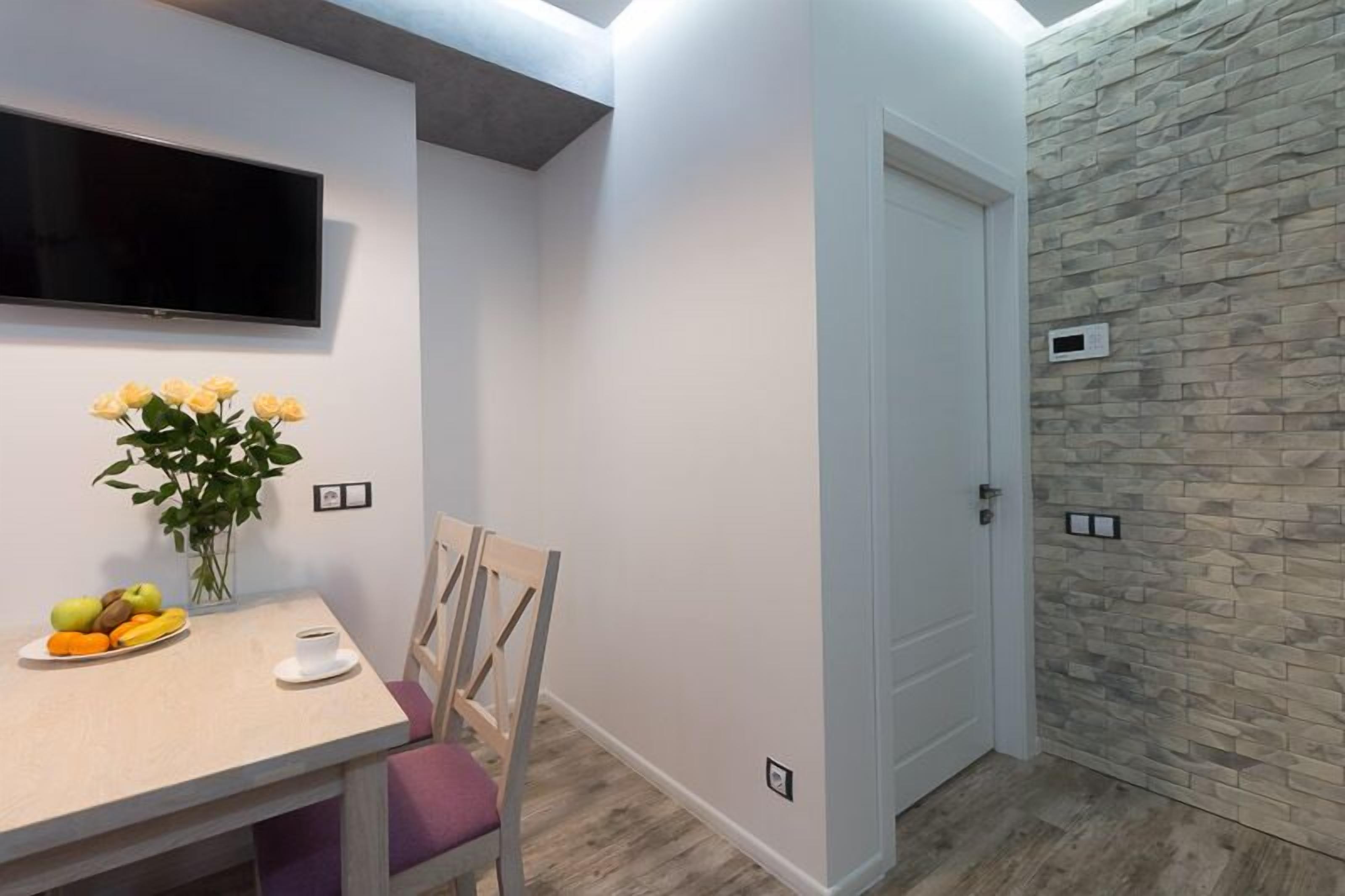 A modern dining area with a table set for two, a vase of yellow flowers, and a flat-screen TV mounted on the wall.