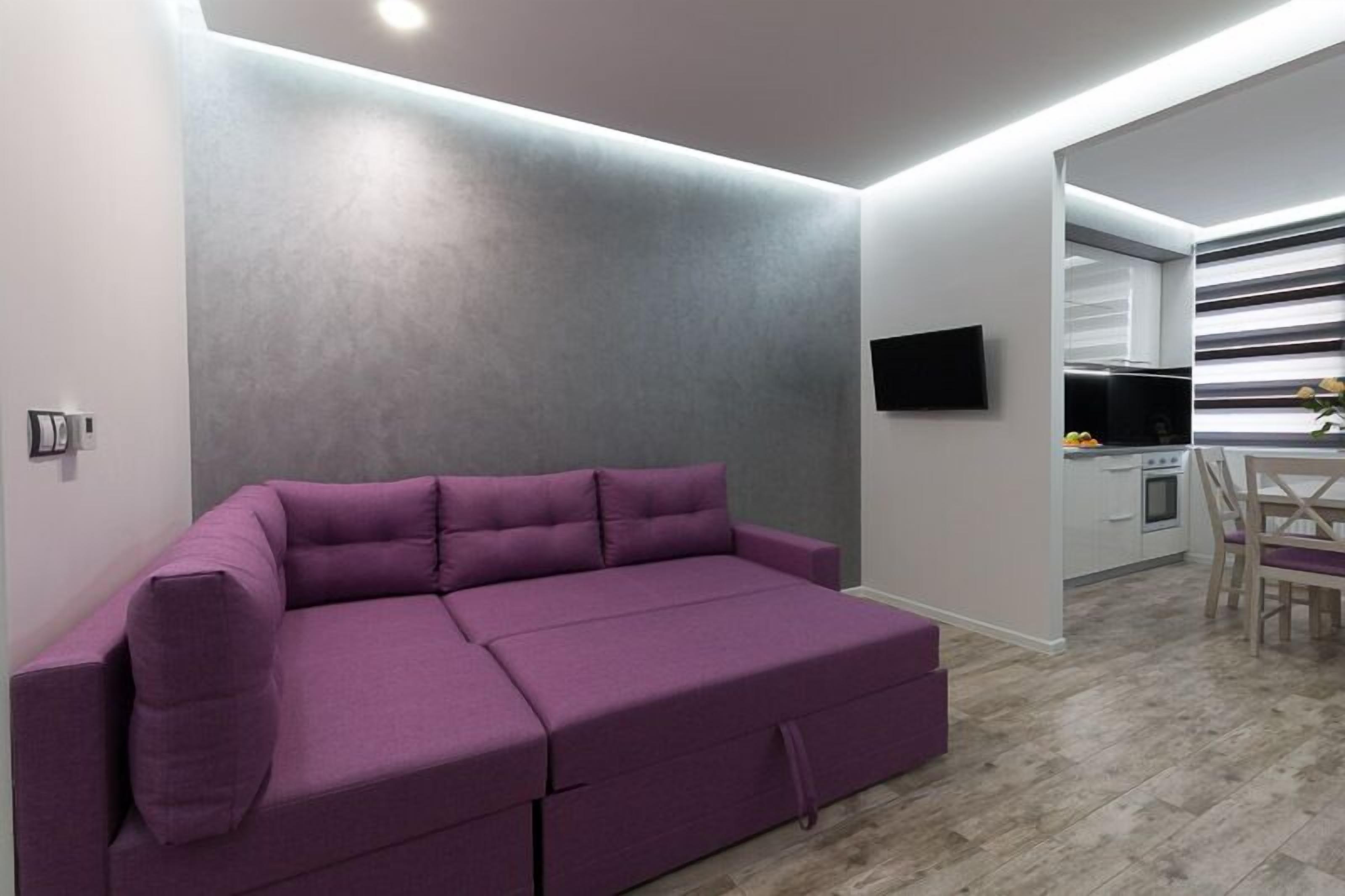 Una sala moderna con un sofá púrpura, un televisor de pantalla plana montado en la pared y una cocina visible a través de un vano abierto.