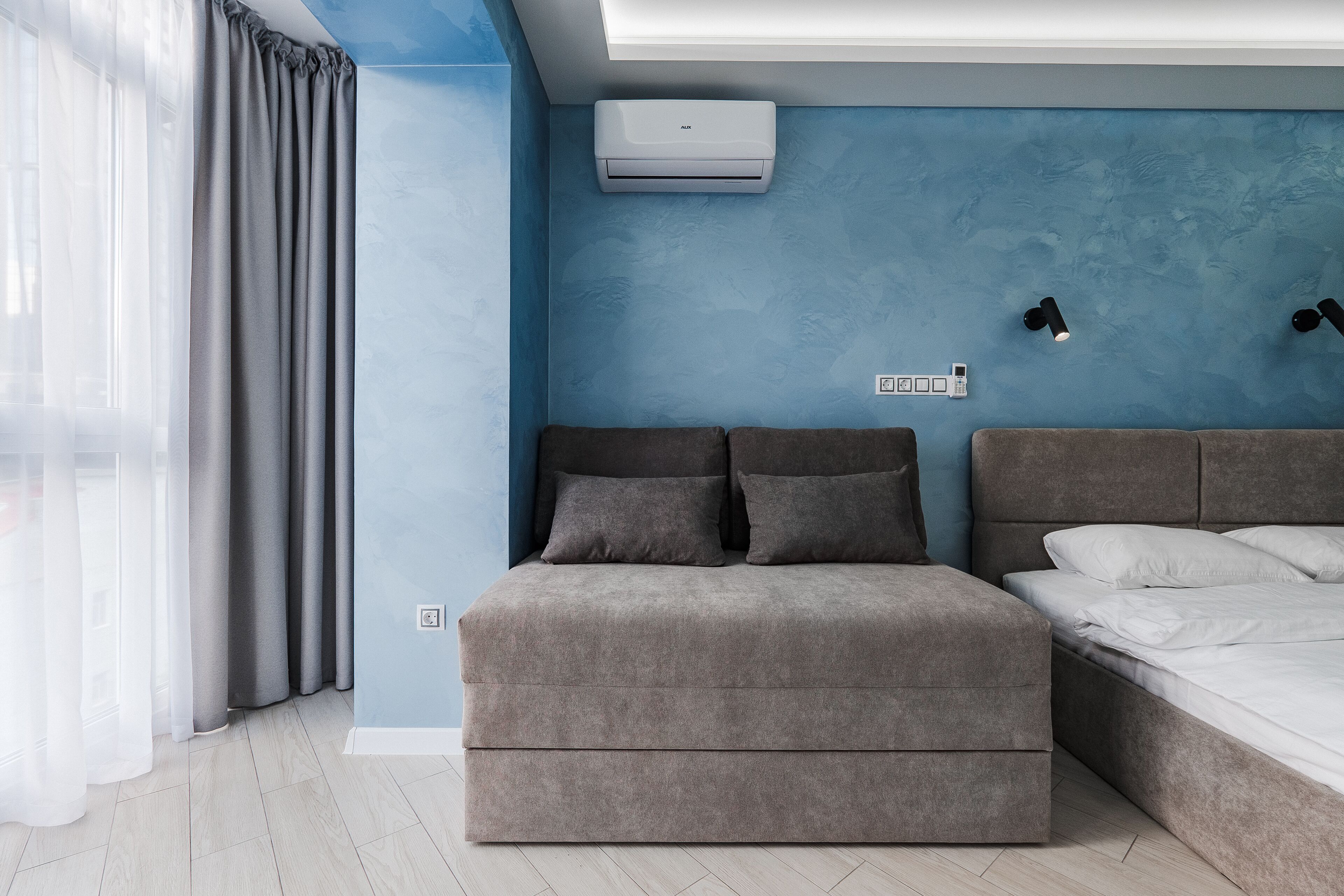A modern bedroom with a blue accent wall, a grey sofa, a bed with white linens, and an air conditioning unit.