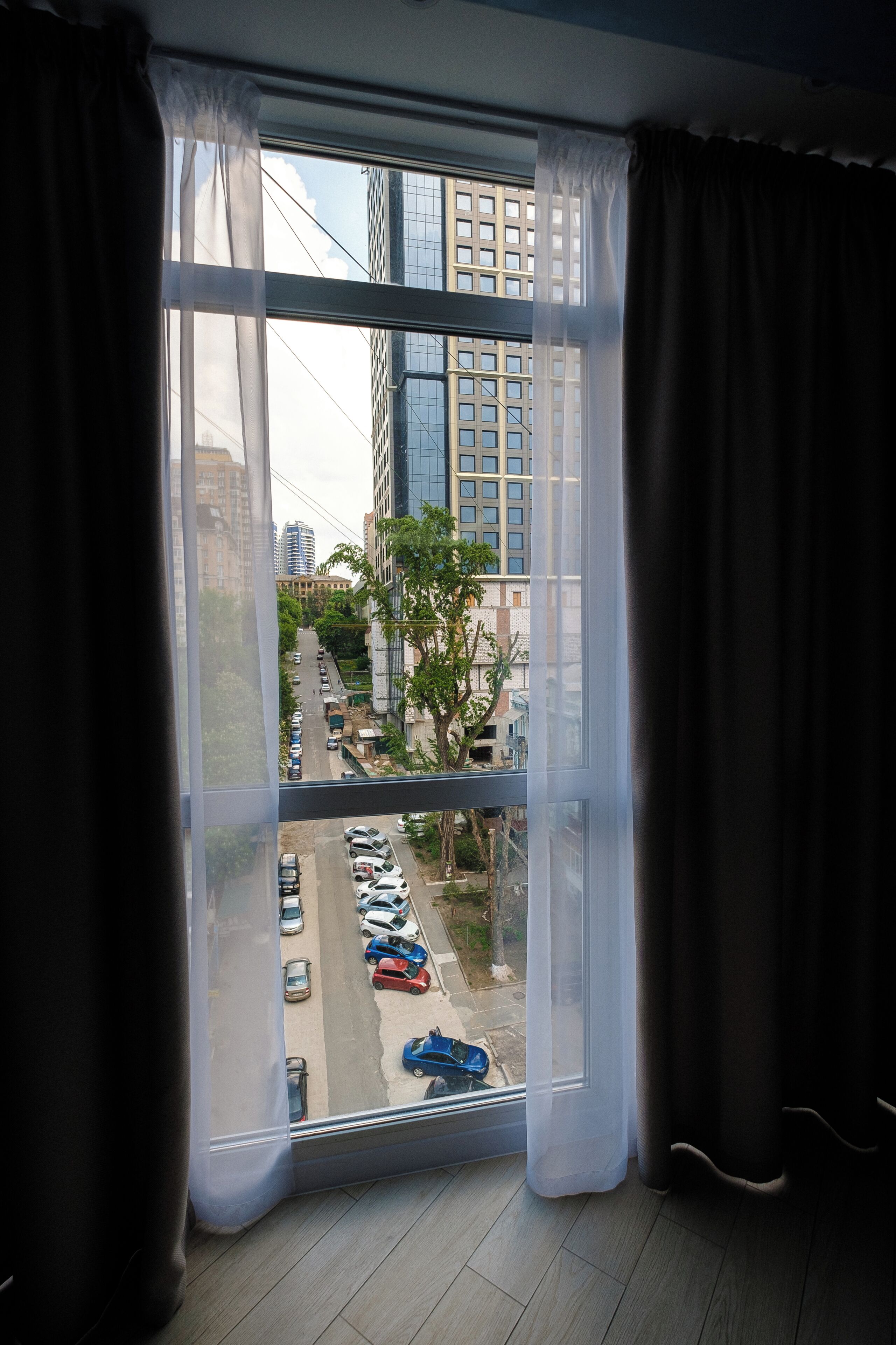 A room with a large window, curtains, and a view of a parking lot and buildings.