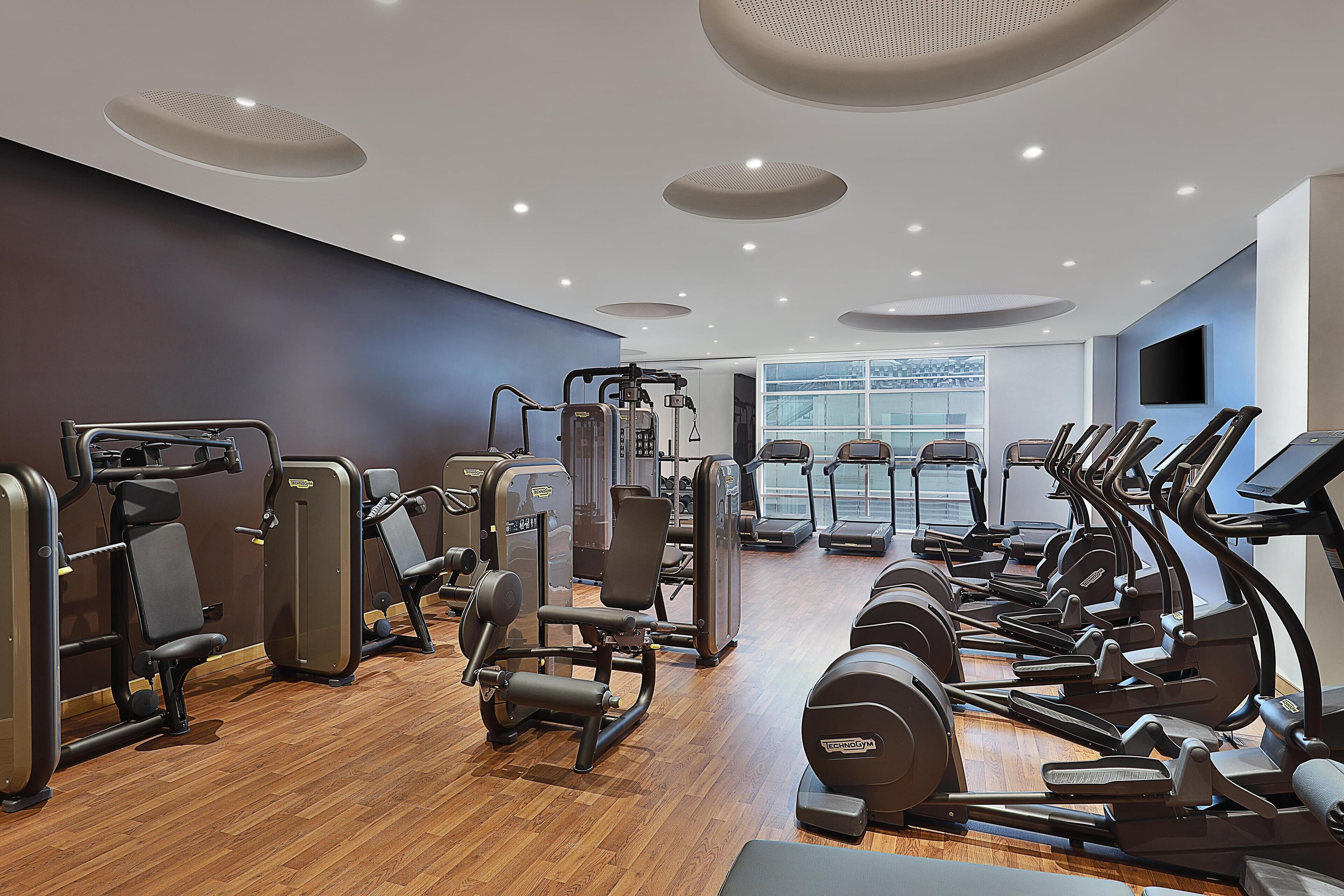 A modern gym with various exercise equipment, large windows, and a wooden floor.