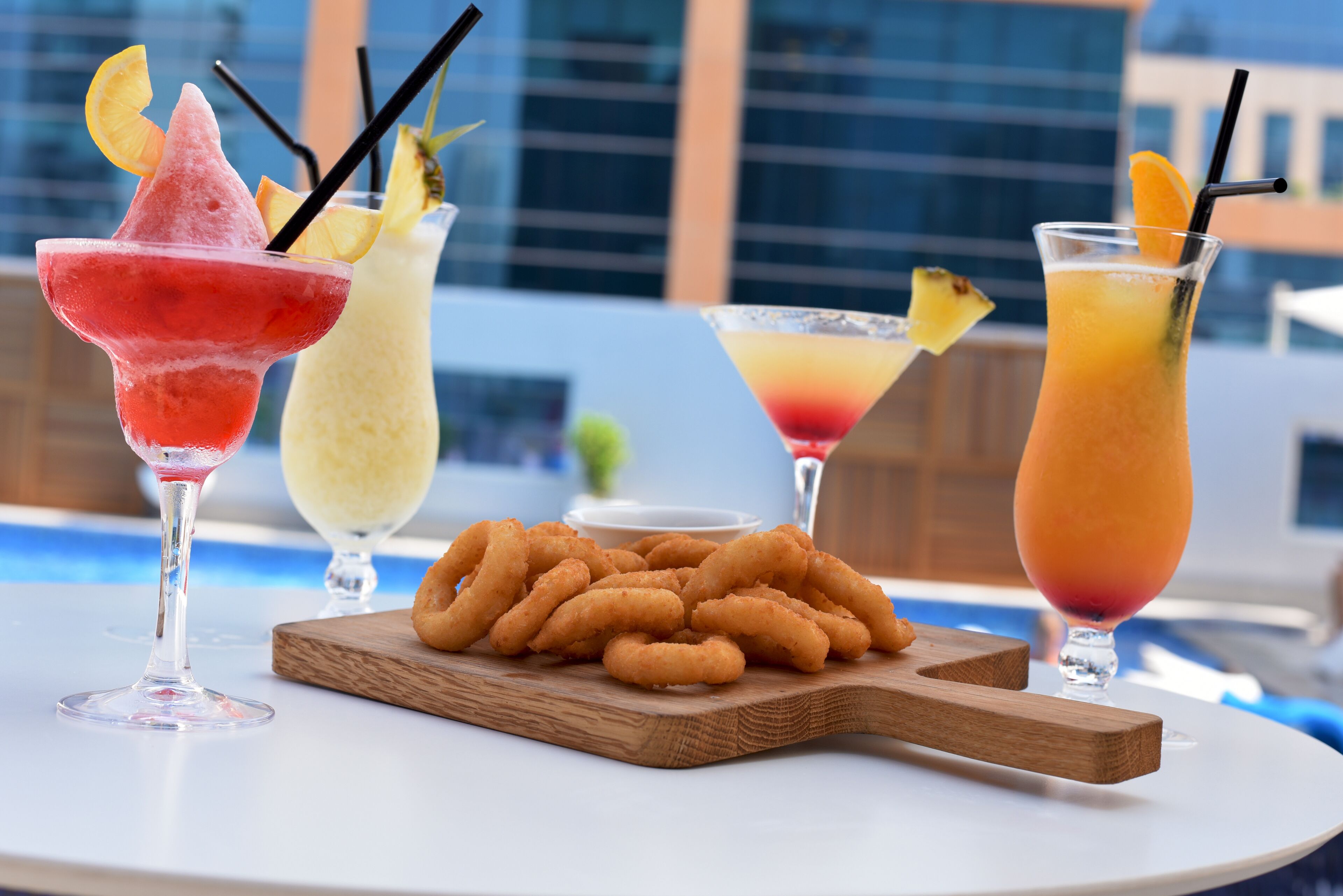 A table with three drinks, a wooden board with fried snacks, and a pool in the background.