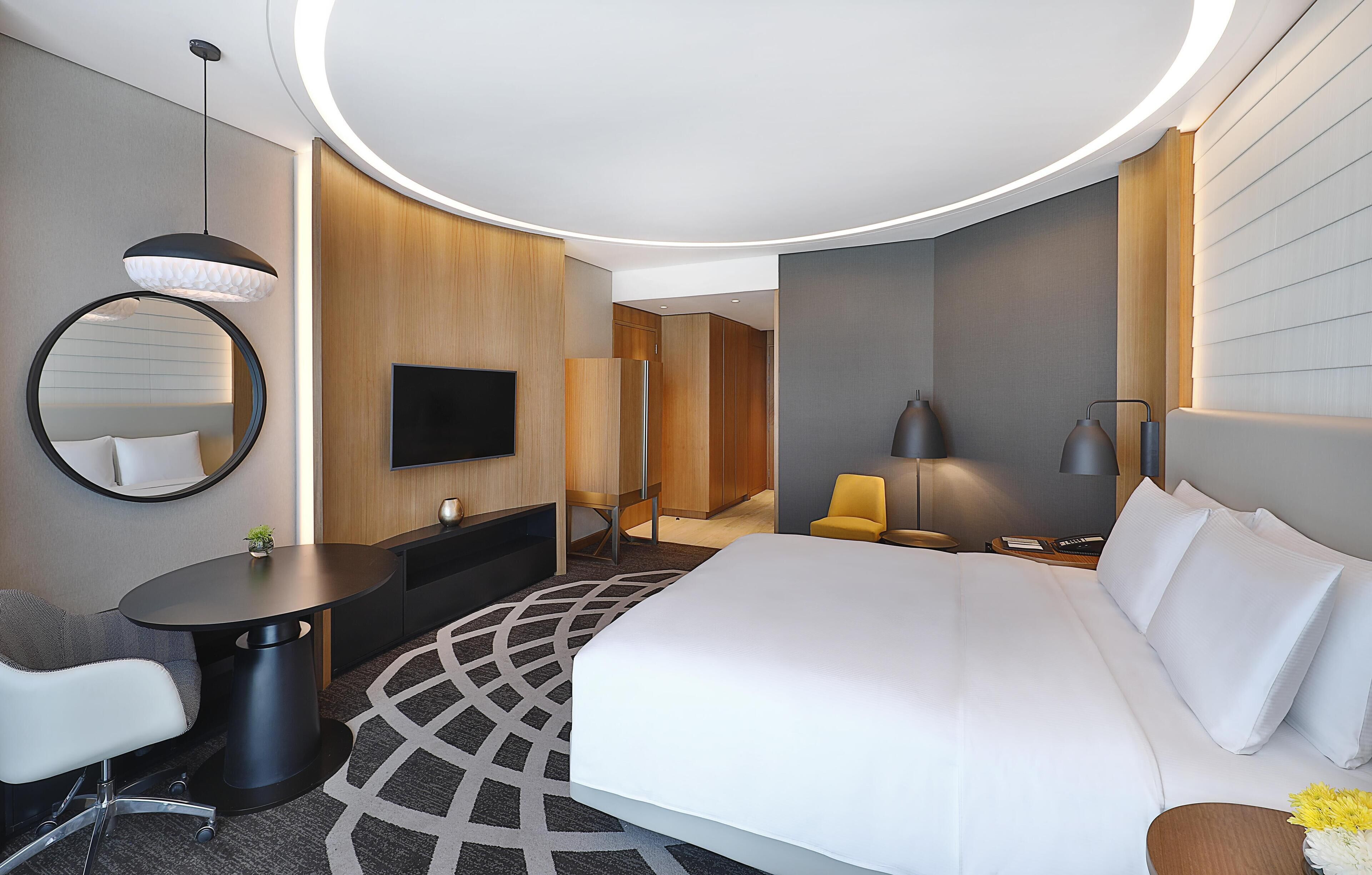 Ein modernes Hotelzimmer mit einem großen Bett, einem Schreibtisch, einem Sessel, einem Fernseher und einem runden Spiegel.