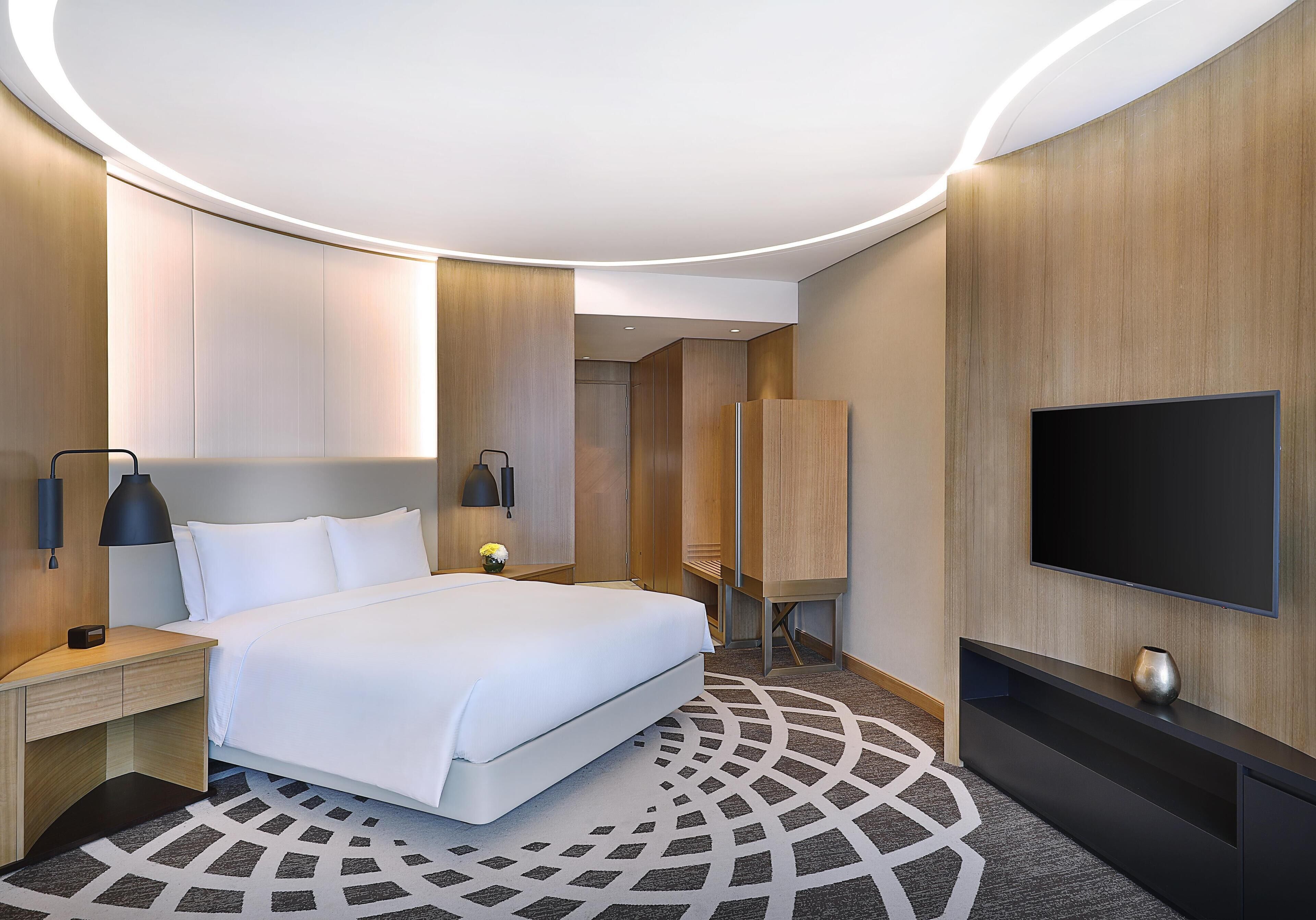 A modern hotel room with a large bed, a flat-screen TV, a desk with a lamp, and a patterned carpet.