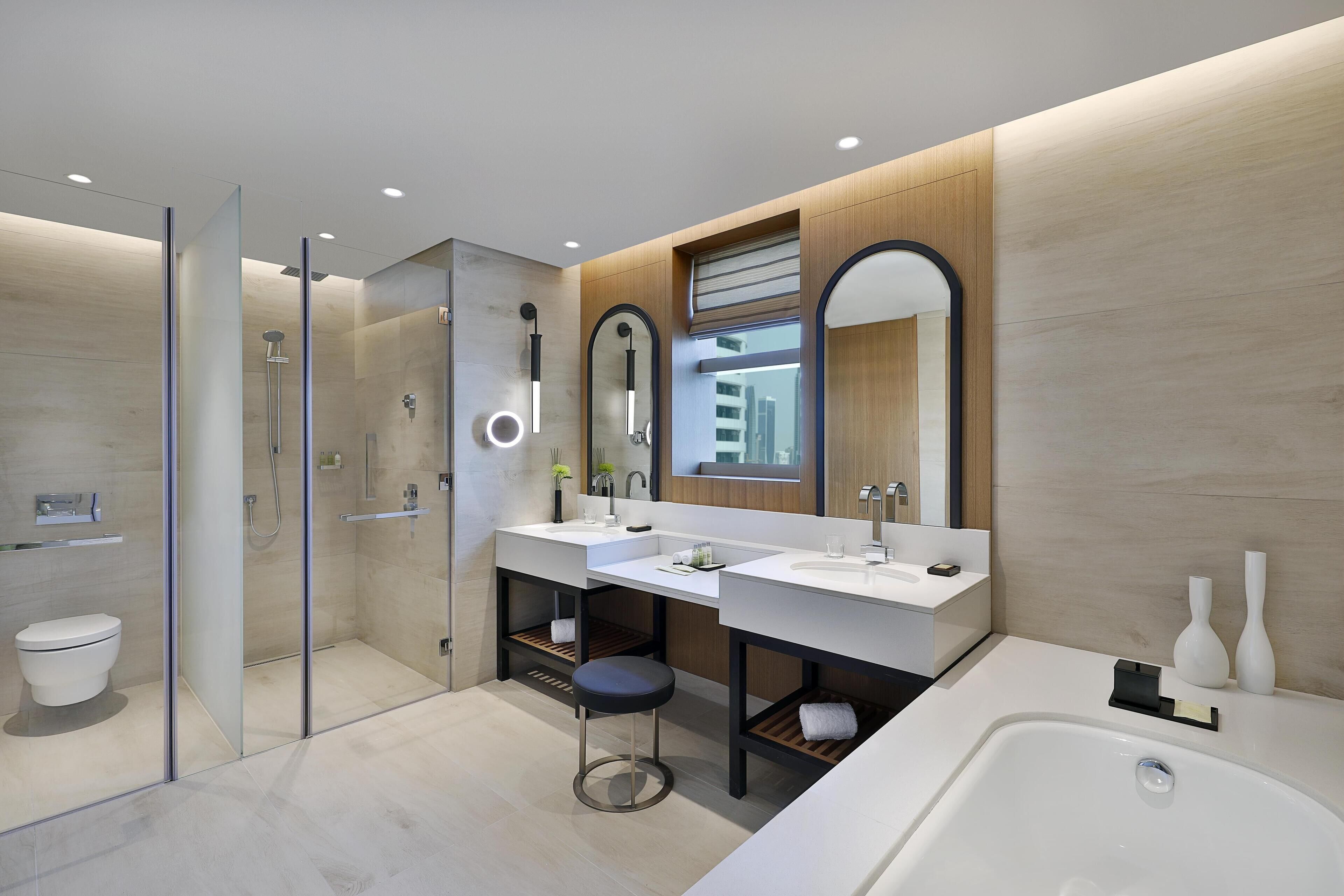 A modern bathroom with a glass-enclosed shower, two sinks, a large bathtub, and a mirror with a wooden panel design.