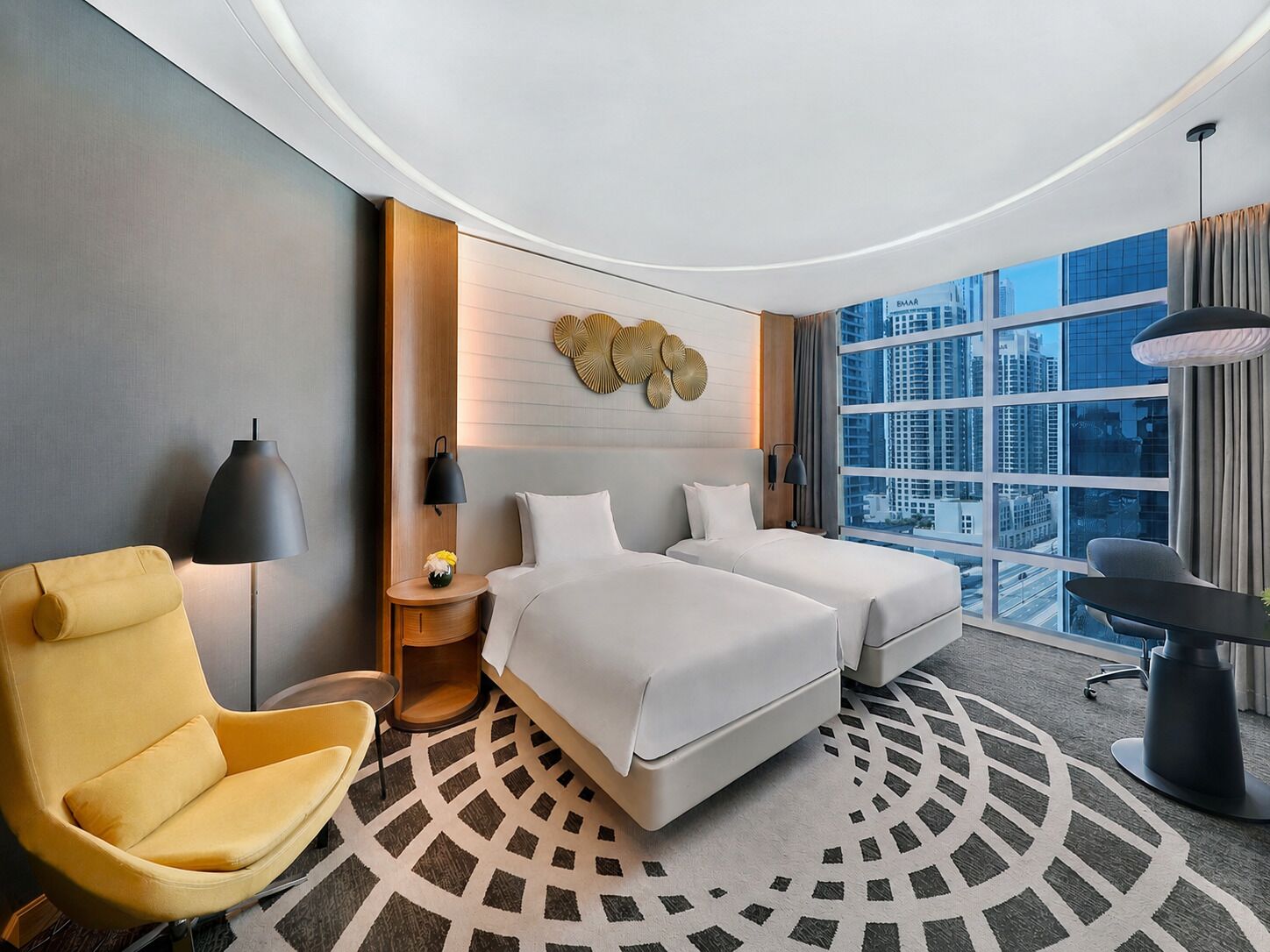 Een hotelkamer met twee bedden, een gele fauteuil, een rond tapijt en een groot raam met uitzicht op de stad.