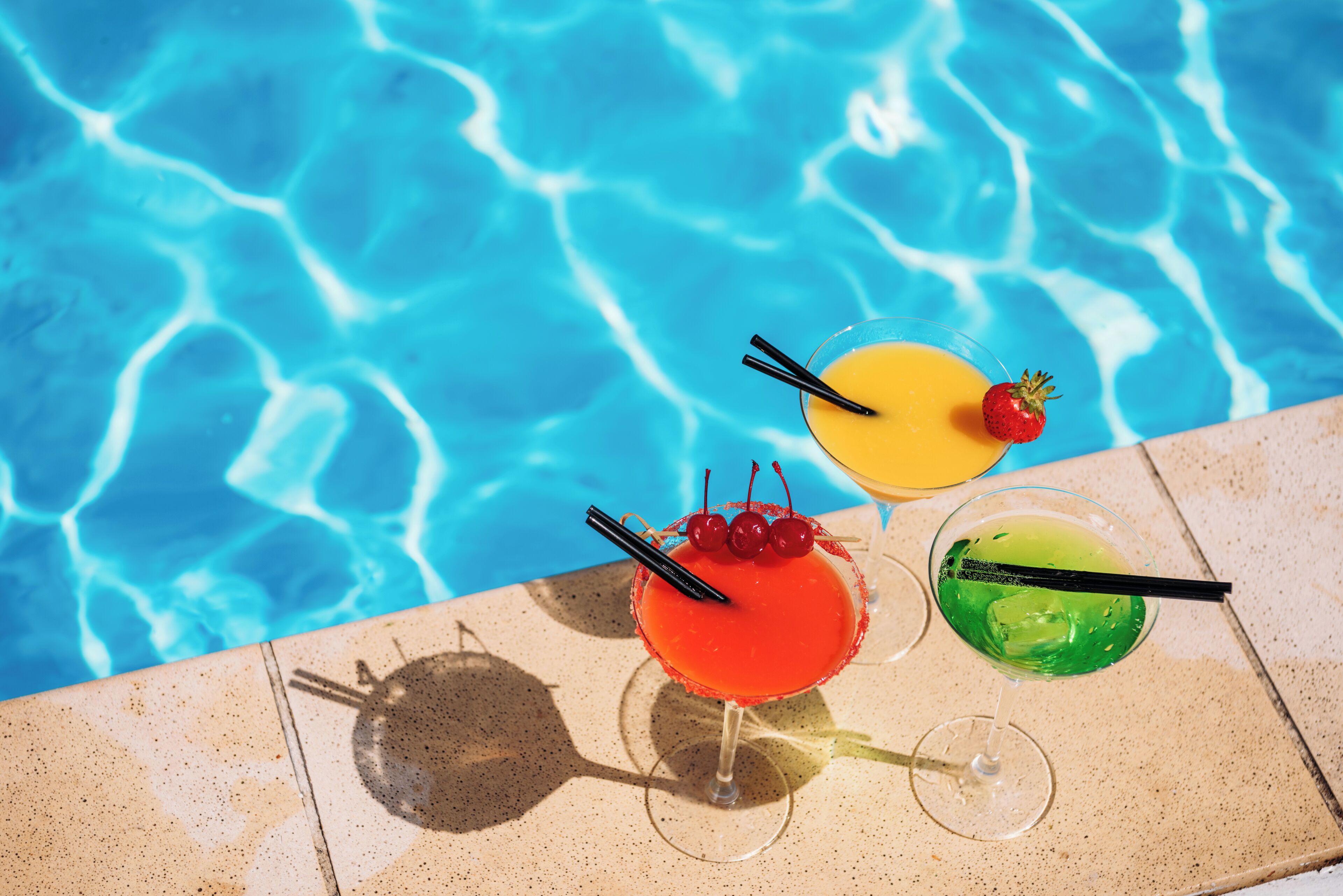 Three cocktails on a poolside table with a clear blue pool in the background.