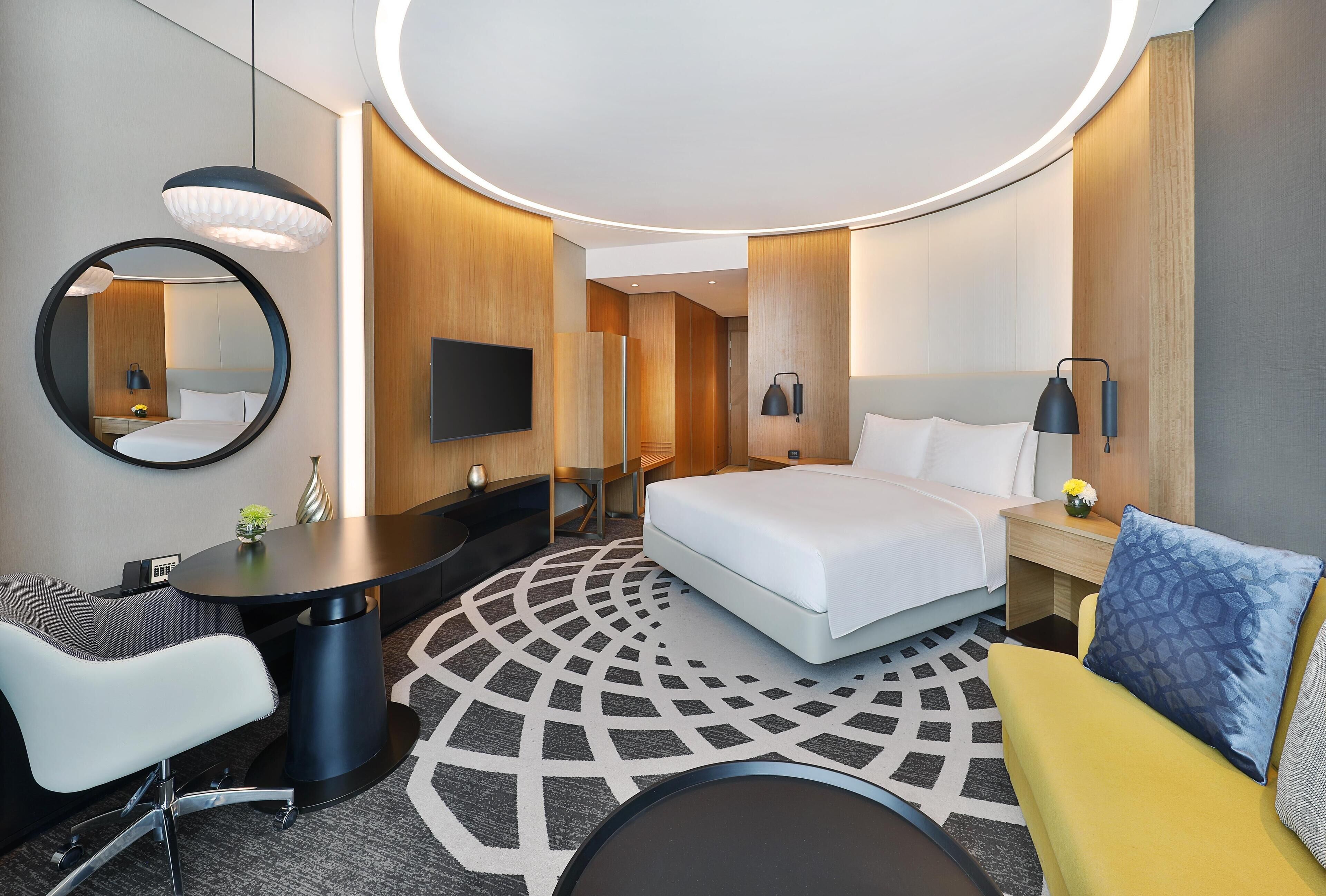 A modern hotel room with a round bed, a desk, a chair, a round table, a mirror, and a TV.