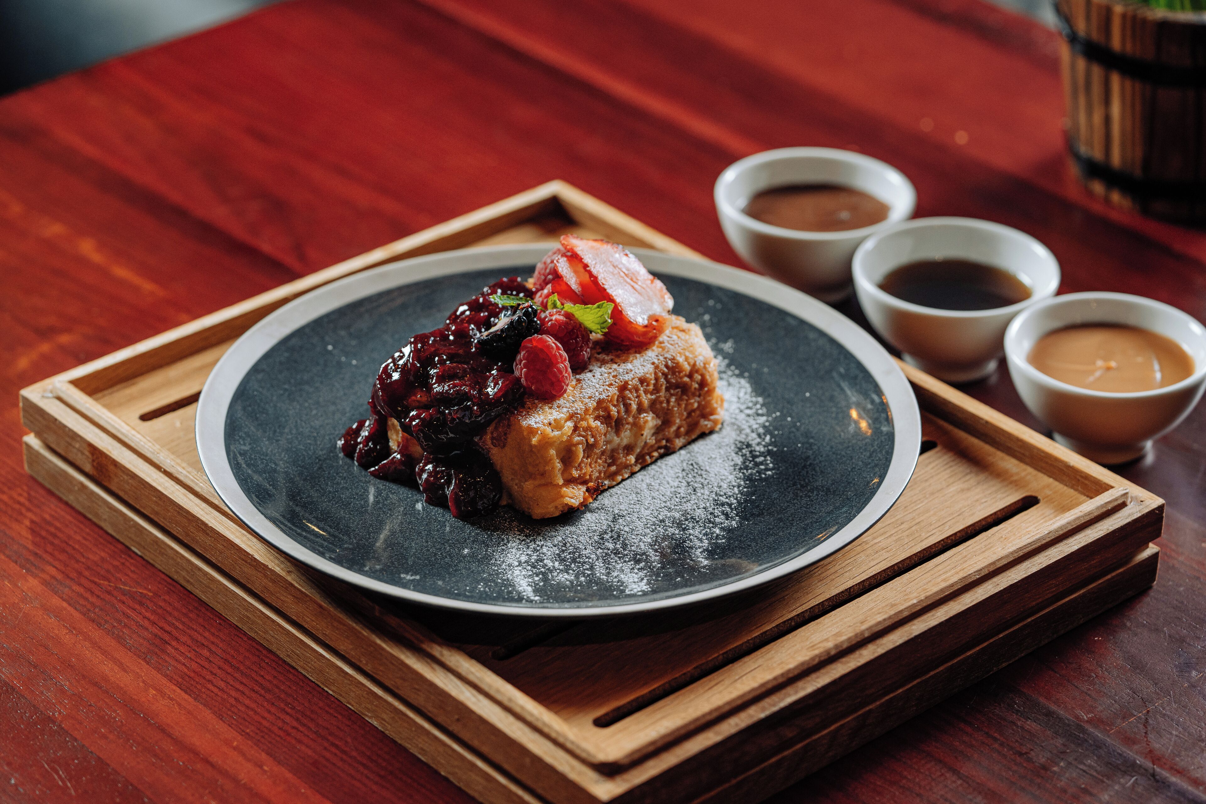 Ein Dessertteller mit einem Kuchenstück, Beeren und Puderzucker auf dunklem Untertaler, dazu drei kleine Soßenbecher auf einer Holzschale.