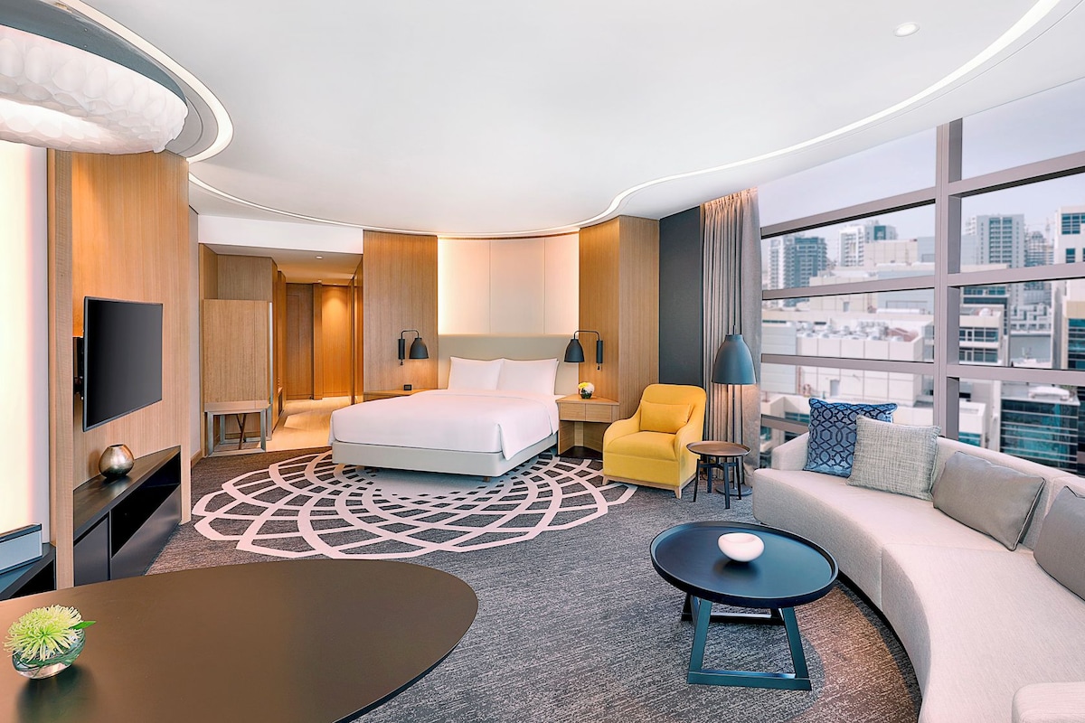 A hotel room with a bed, a sofa, a small table, a TV, and a view of the city.