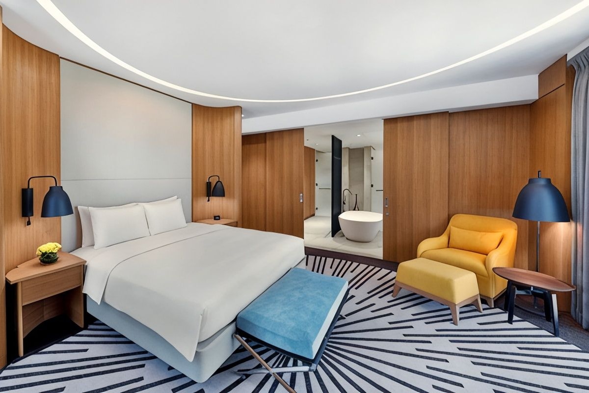 A modern hotel room with a large bed, a desk, a chair, and a small table.