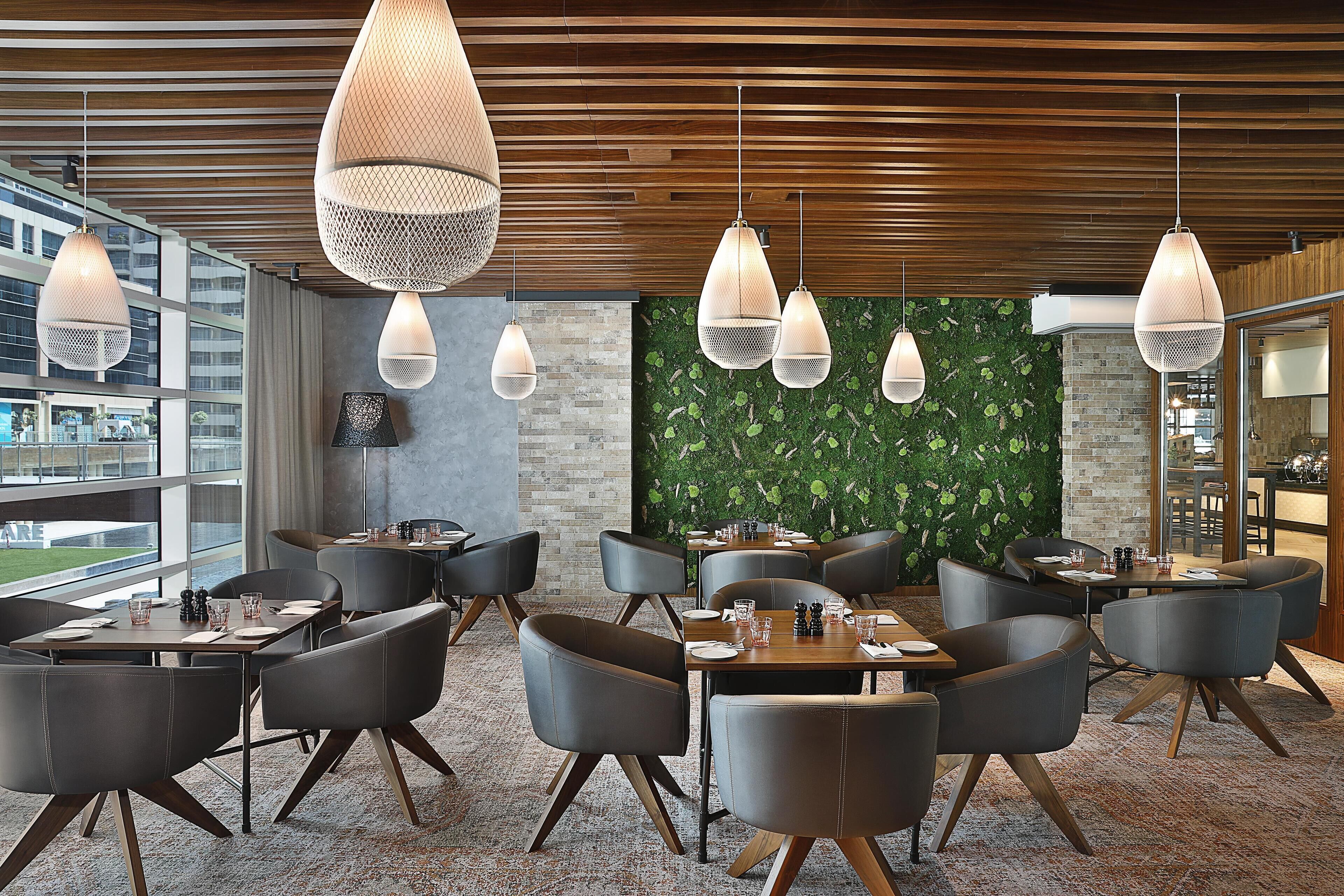 A modern dining area with wooden ceiling, hanging pendant lights, and a green wall with foliage. Tables and chairs are arranged for dining.