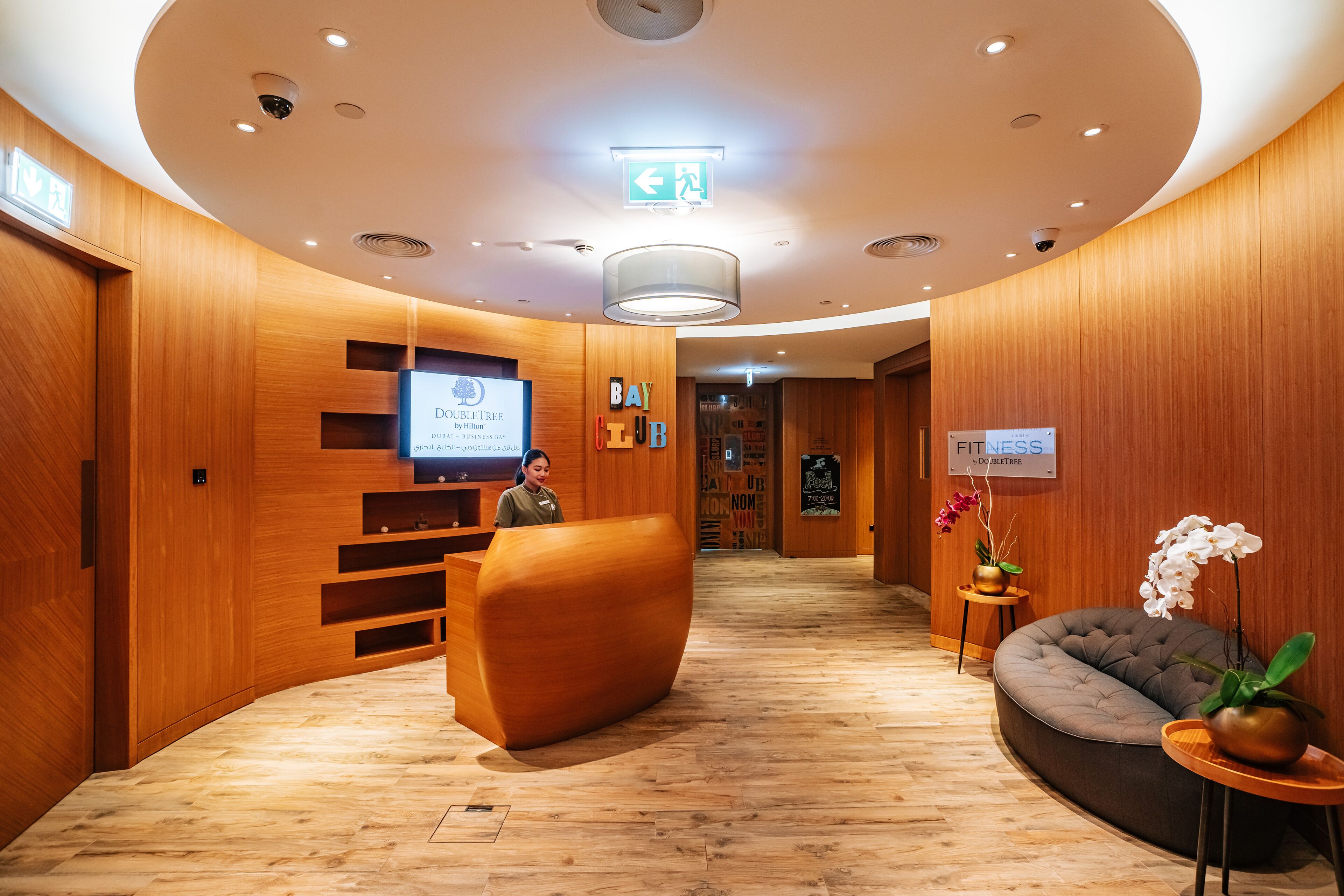 Un hall d'accueil d'hôtel avec un décor intérieur en bois, un comptoir d'accueil, un espace salon et un panneau indiquant l'emplacement d'une salle de sport.