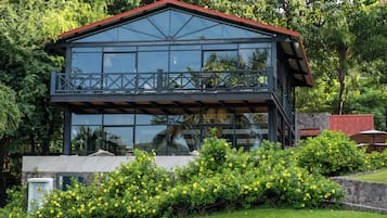 A two-story building with a balcony, surrounded by greenery and yellow flowers.
