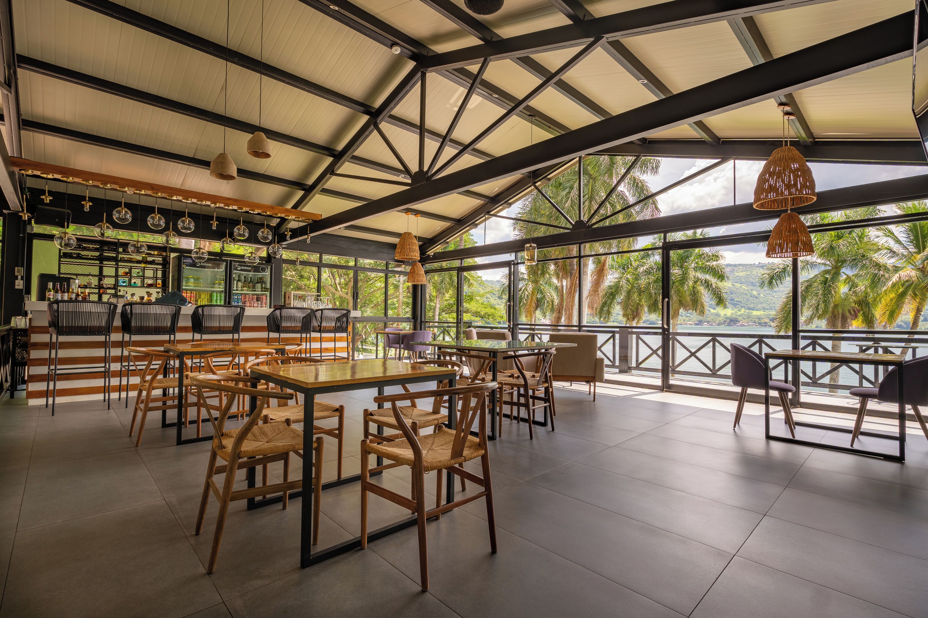 Un espacio al aire libre con mesas y sillas, un bar con mostrador refrigerado, y vistas a palmeras y una masa de agua.