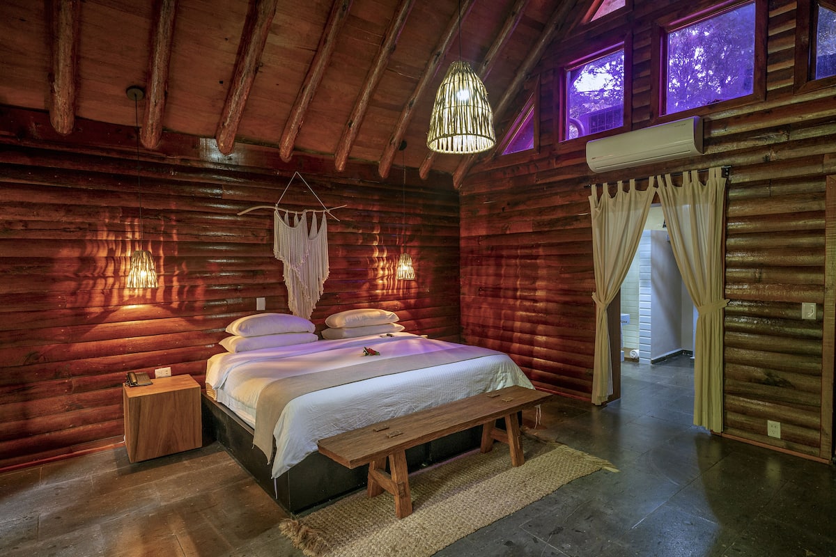 A log cabin bedroom with a wooden bed, bedside lamps, and a bench.