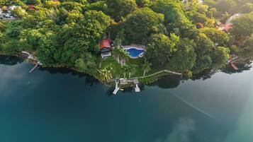 An aerial view of a tropical island with a swimming pool, a dock, and several buildings surrounded by dense greenery.