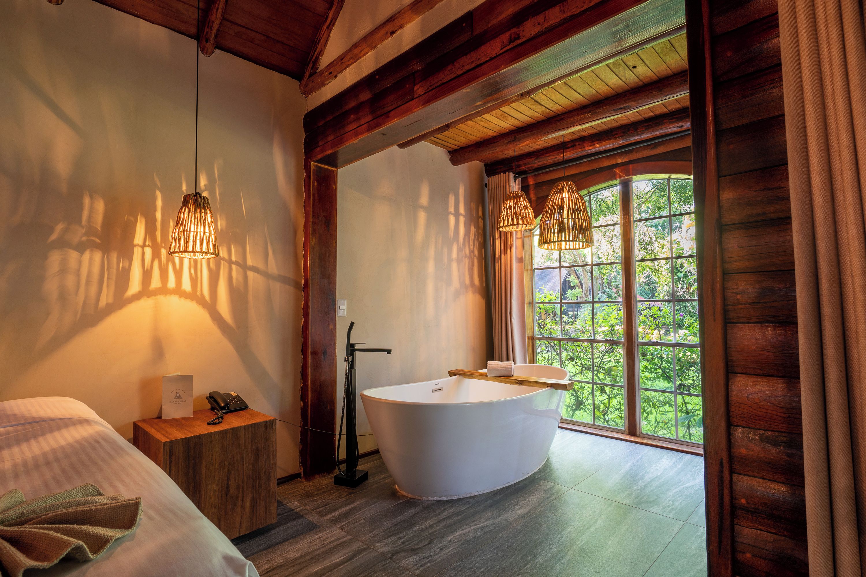 A modern bathroom with a freestanding bathtub, wooden beams, and a large window with a view of greenery.