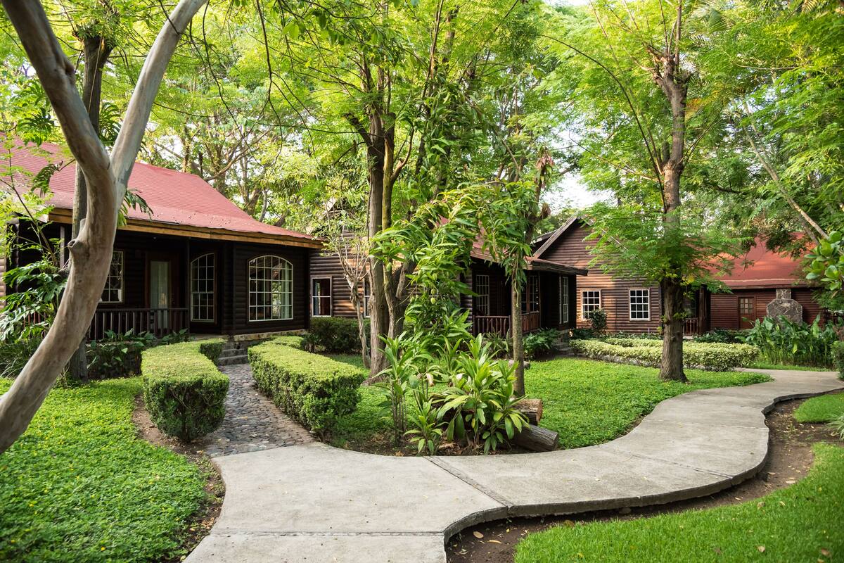 A wooden house with a red roof, surrounded by lush greenery and a curved concrete pathway.
