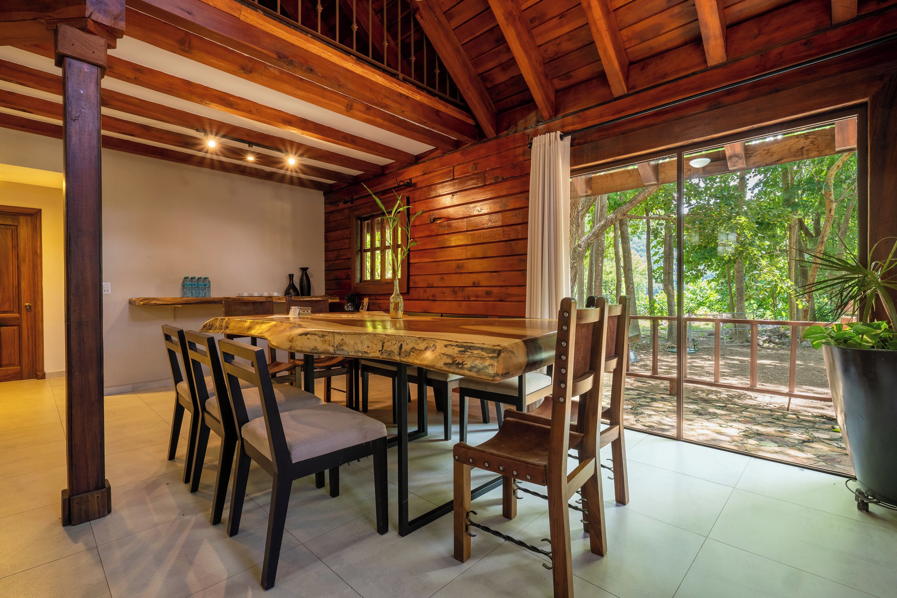 Uma área de refeições com mesa e cadeiras de madeira, janela com cortinas e vista para o verde lá fora.