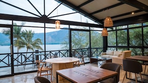 A spacious indoor area with large windows overlooking a body of water and mountains. There are multiple seating arrangements including tables and chairs. - Santa María Resort (La Laguna)