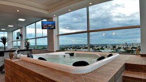 Un espace spa moderne doté d’un grand jacuzzi, de larges fenêtres offrant une vue sur la ville et d’un téléviseur à écran plat fixé au mur.