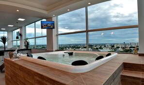 Een moderne spa-ruimte met een ruime jacuzzi, grote ramen met uitzicht op de stad en een flatscreen televisie aan de muur.