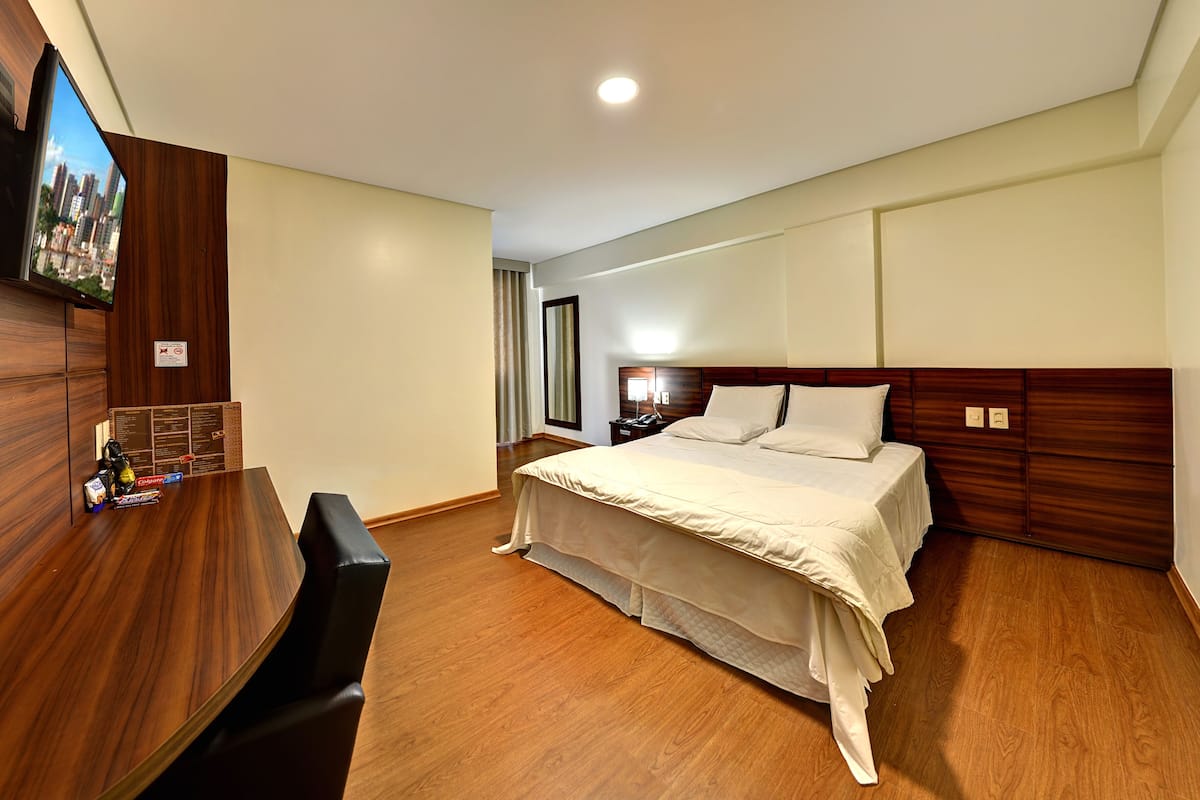 Une chambre d’hôtel avec un lit en bois, un bureau avec une chaise, un téléviseur à écran plat fixé au mur et une vue sur la ville.