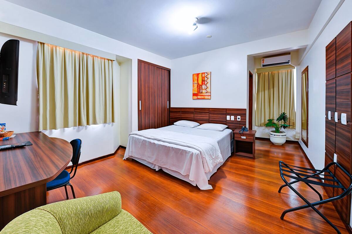 Une chambre d’hôtel avec un plancher en bois, un lit avec du linge de maison blanc, un bureau avec une chaise, une plante en pot et un tableau accroché au mur.