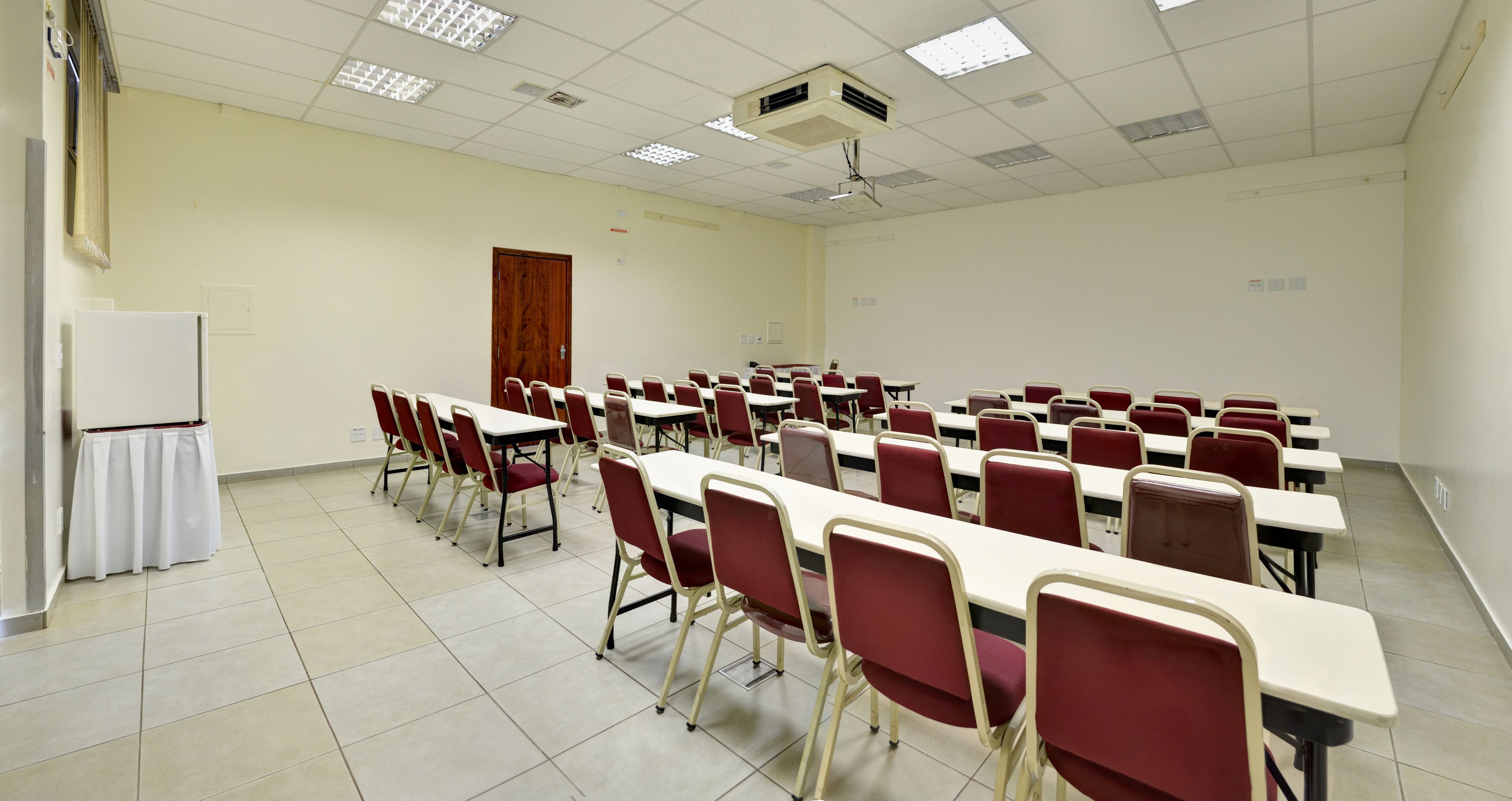 Uma sala de conferências com fileiras de mesas e cadeiras, um quadro branco e um projetor no teto.