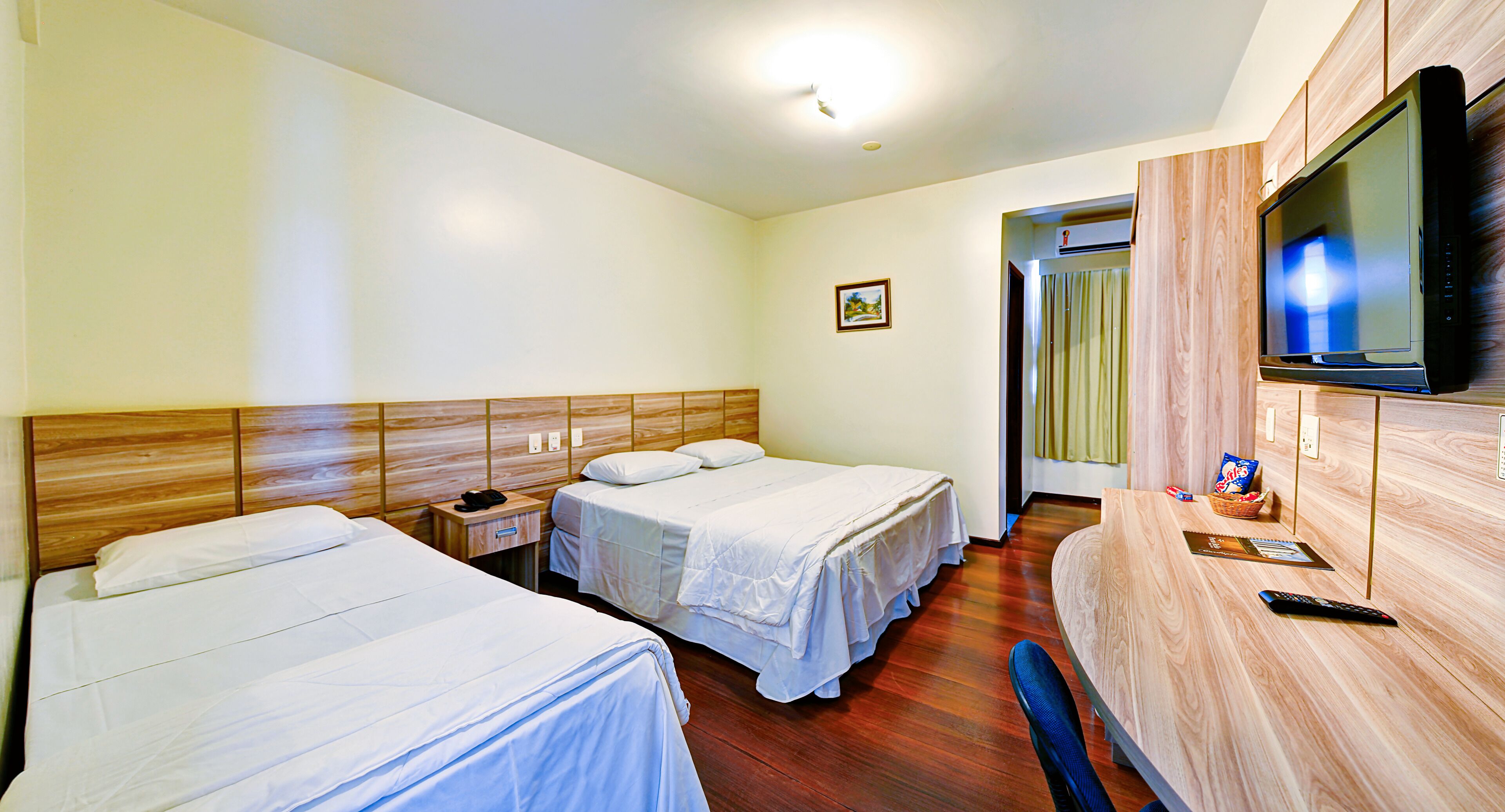 Quarto de hotel com duas camas, uma escrivaninha com televisão e cabeceira de madeira.