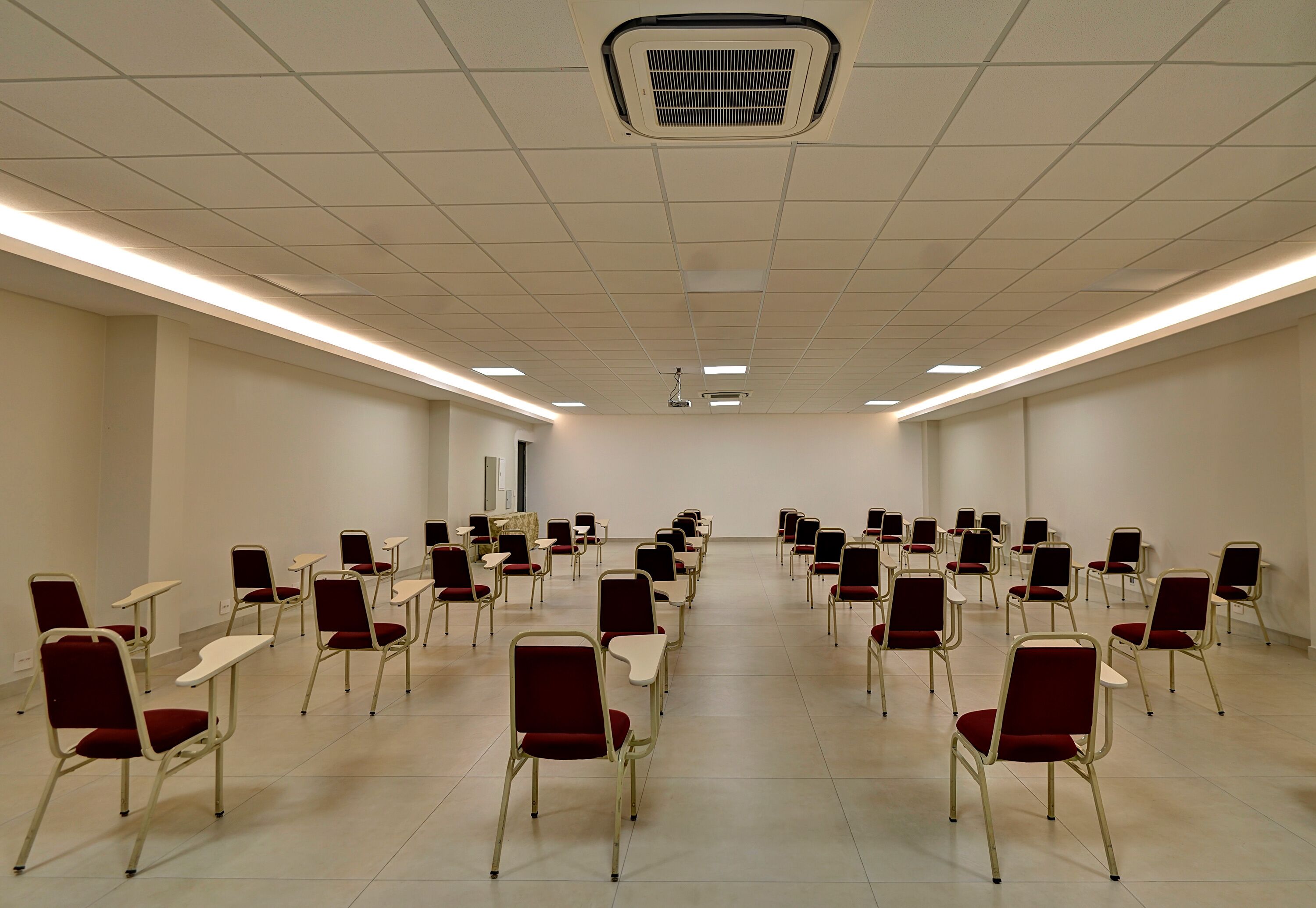 Un salón de conferencias con filas de sillas y mesas, un proyector y una rejilla de ventilación en el techo.