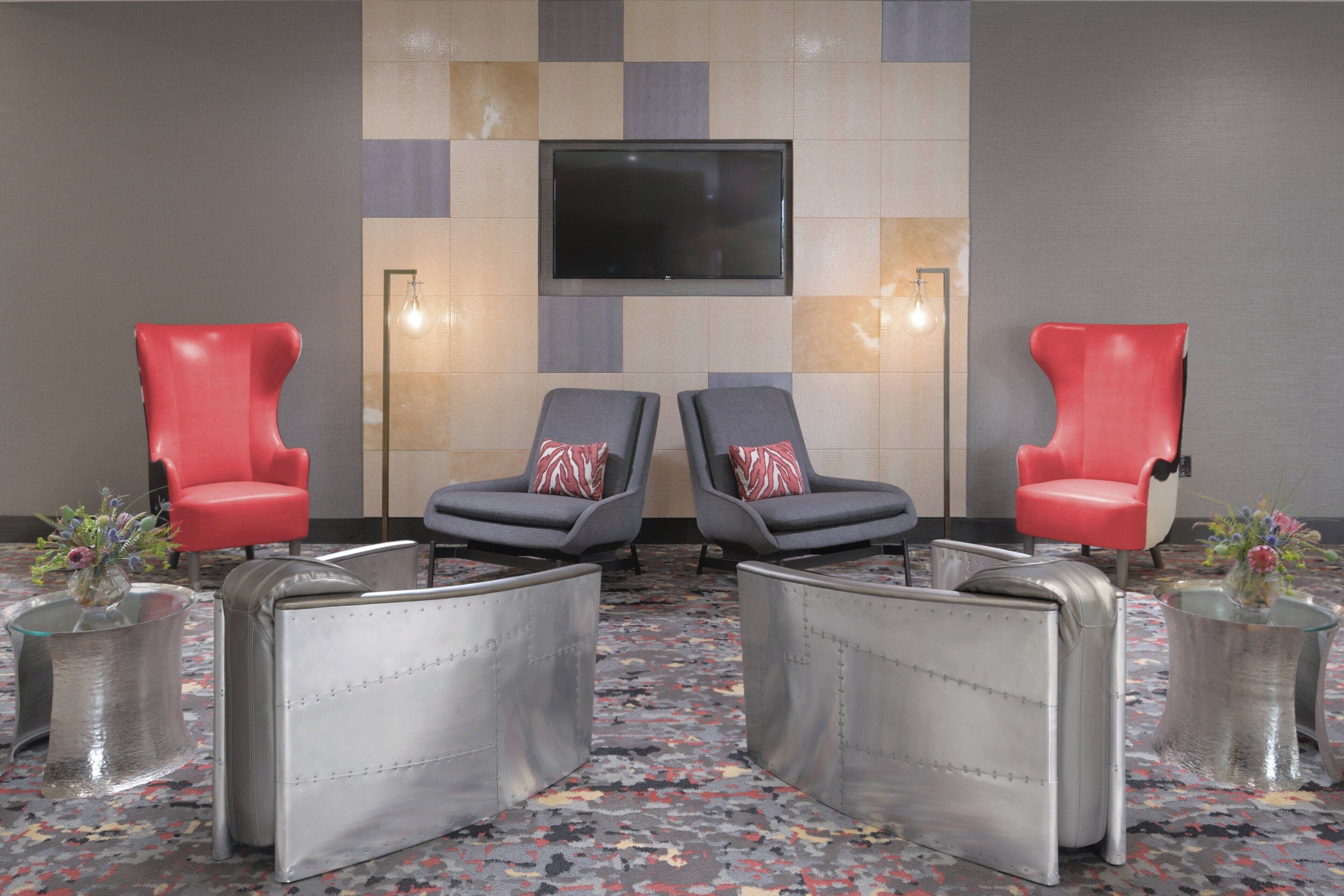 A room with two red chairs, two grey chairs, a TV mounted on the wall, and a patterned carpet.
