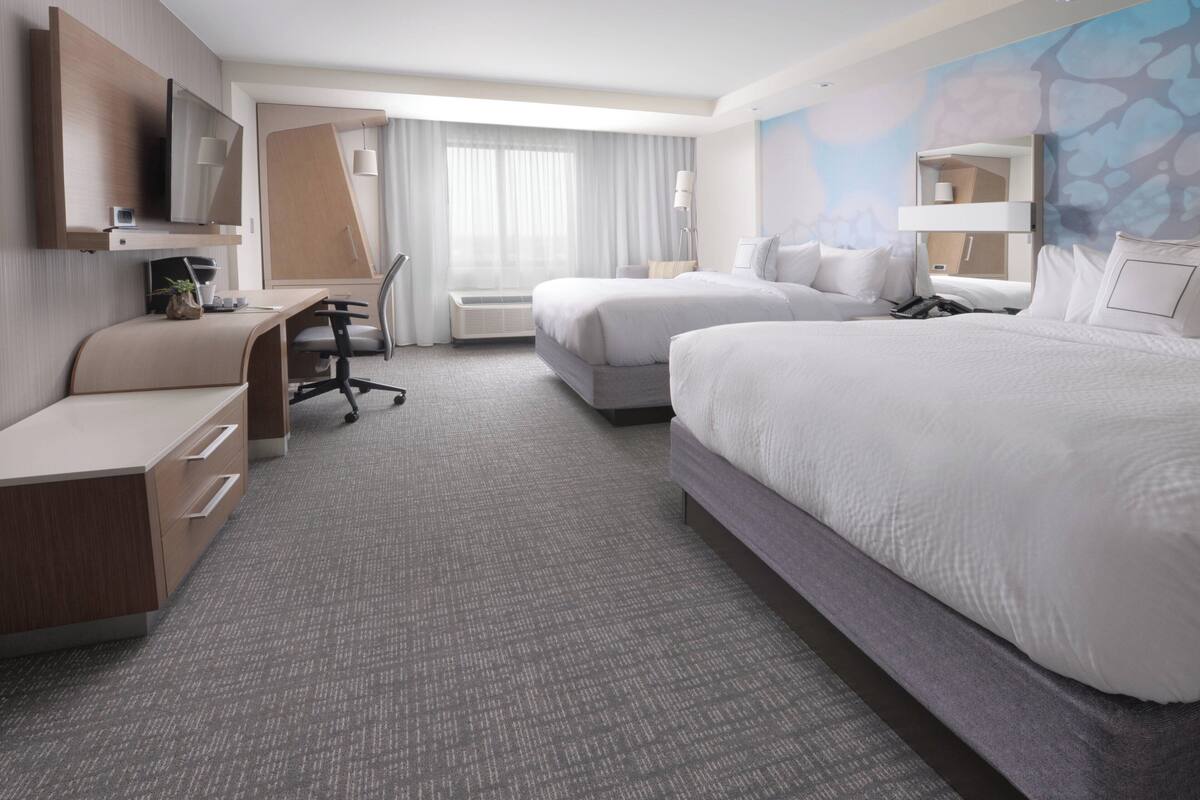 A hotel room with two beds, a desk, a chair, a TV, and a large window with curtains.
