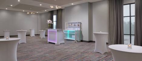 A hotel room with a carpeted floor, white round tables, a reception desk, and a window with curtains.