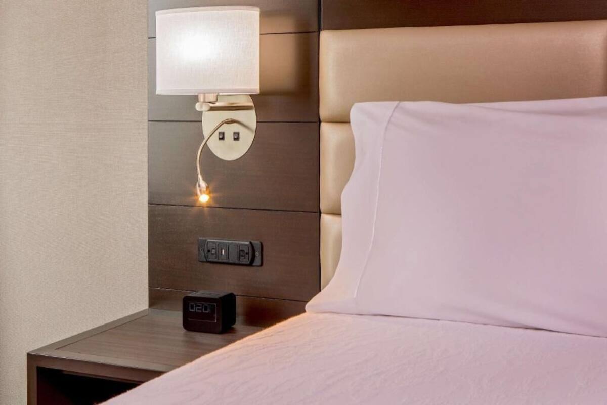 Habitación de hotel con cama, mesita de noche, lámpara de pared y reloj digital.