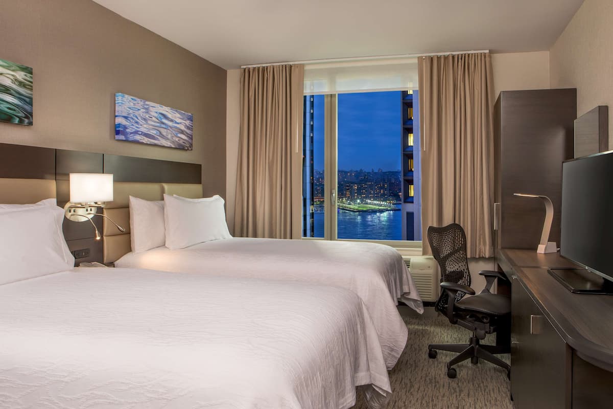 Habitación de hotel con dos camas, un escritorio con televisor, una silla y un ventanal con vista a la ciudad.