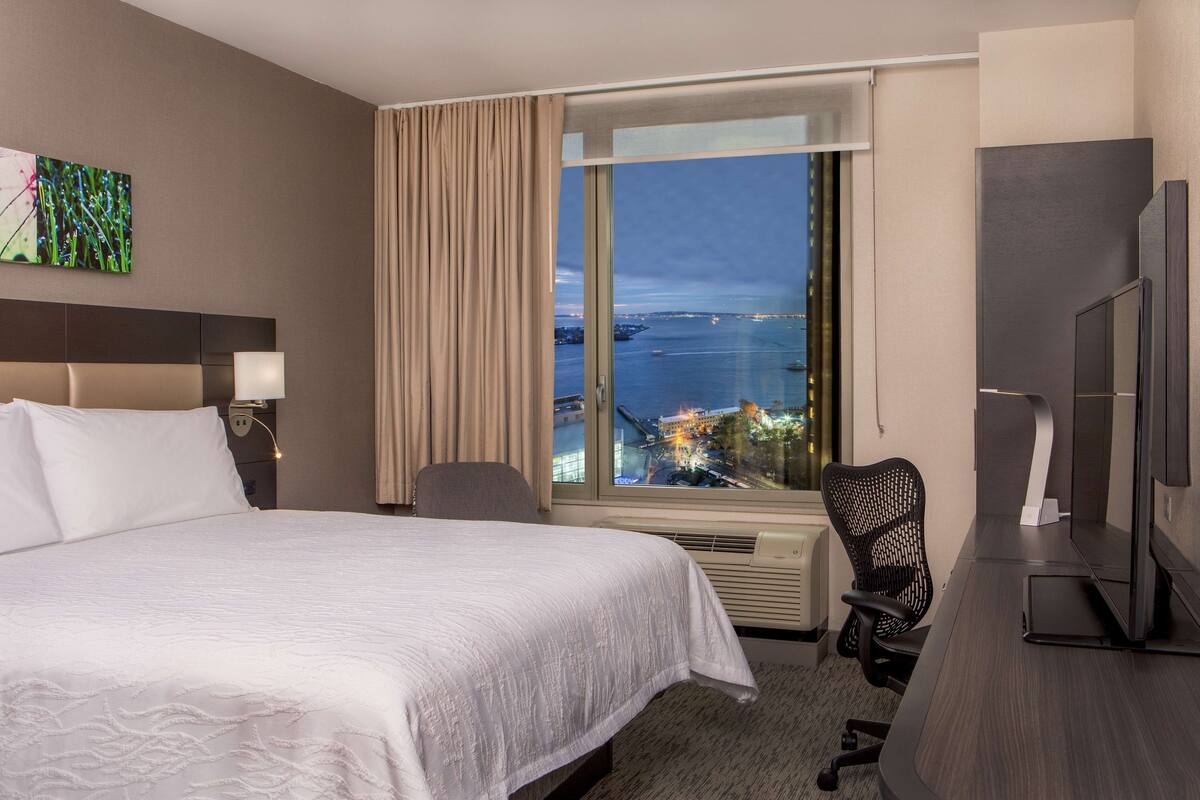 Habitación de hotel con una cama grande, un escritorio con computadora, televisor y ventana con vista a la ciudad.