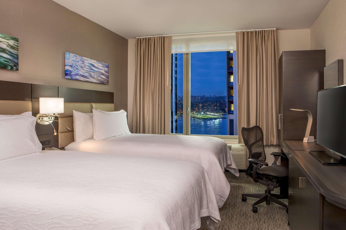 Une chambre d’hôtel avec deux lits, un bureau avec une télévision et une vue sur le paysage urbain de nuit.