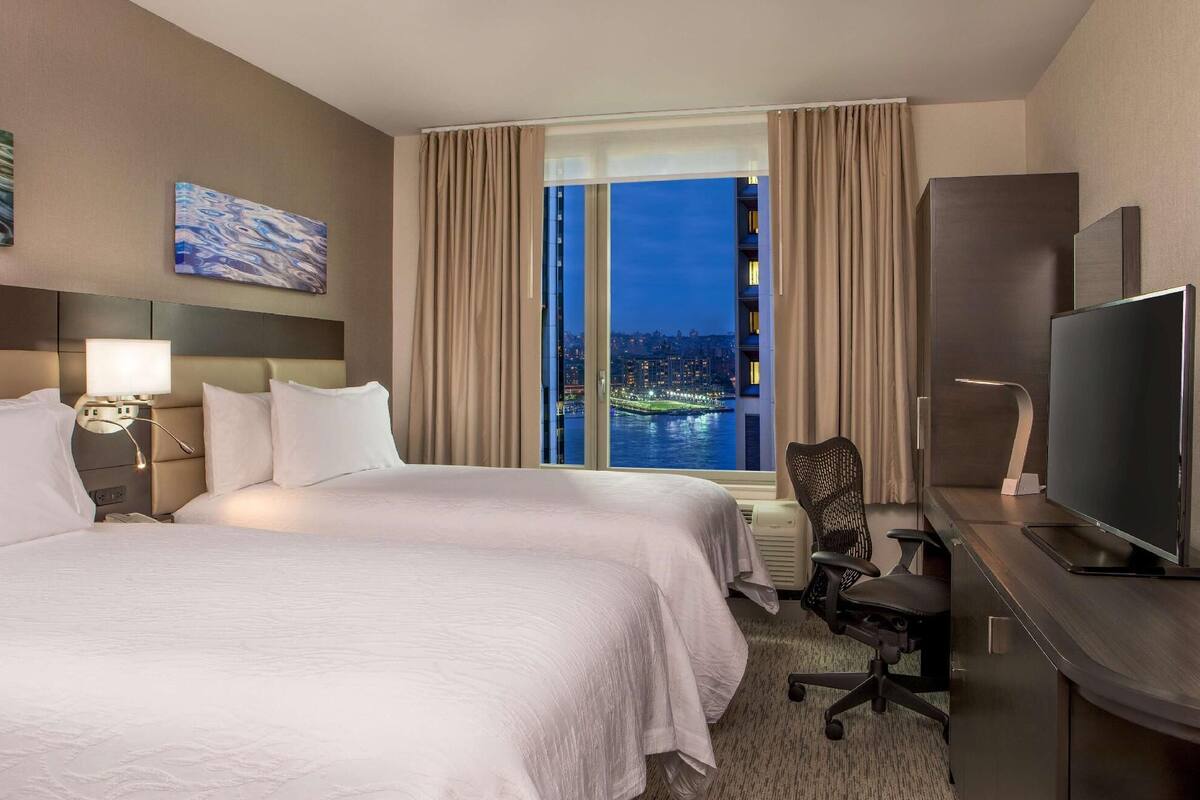 Habitación de hotel con una cama grande, un escritorio con televisión, una silla y vistas a un paisaje urbano nocturno.